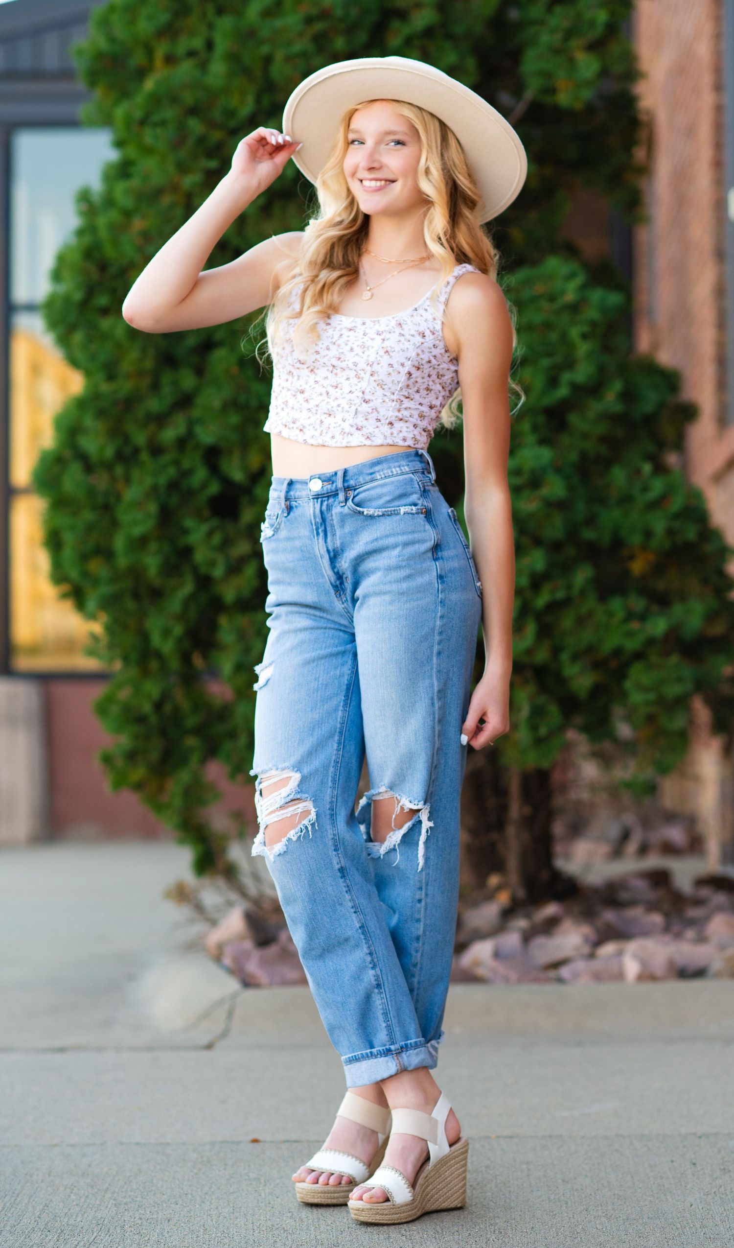 Woman in floral crop top and ripped jeans poses outdoors wearing sun hat and wedge sandals.