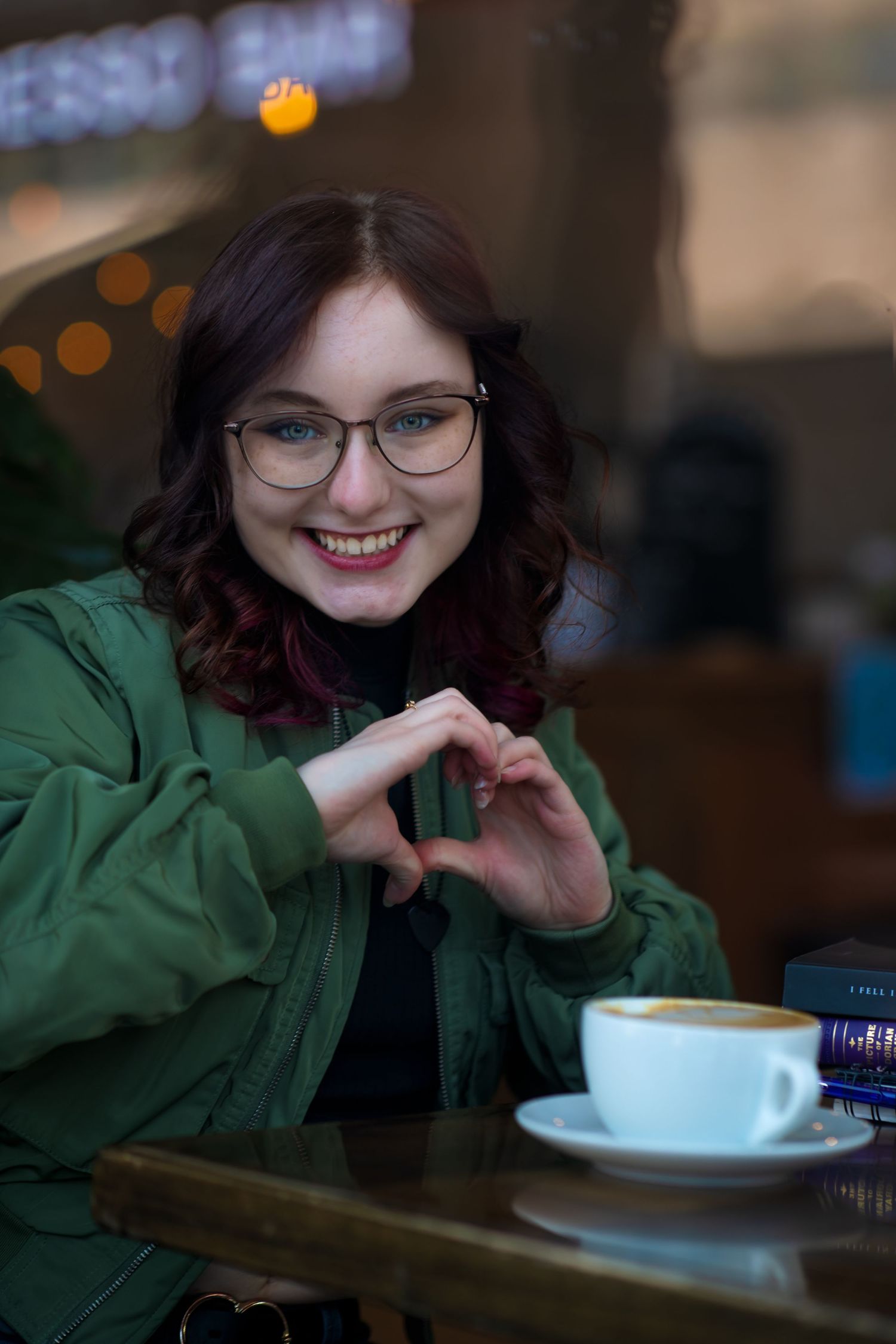Person makes heart shape with hands while sitting at cafe table with coffee cup.
