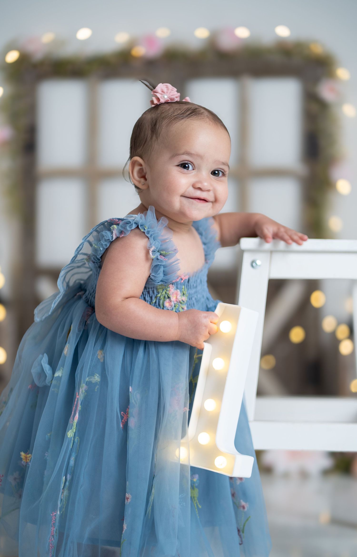 A smiling baby girl in a blue floral tulle dress holds a lit number 1 sign, celebrating her first birthday.