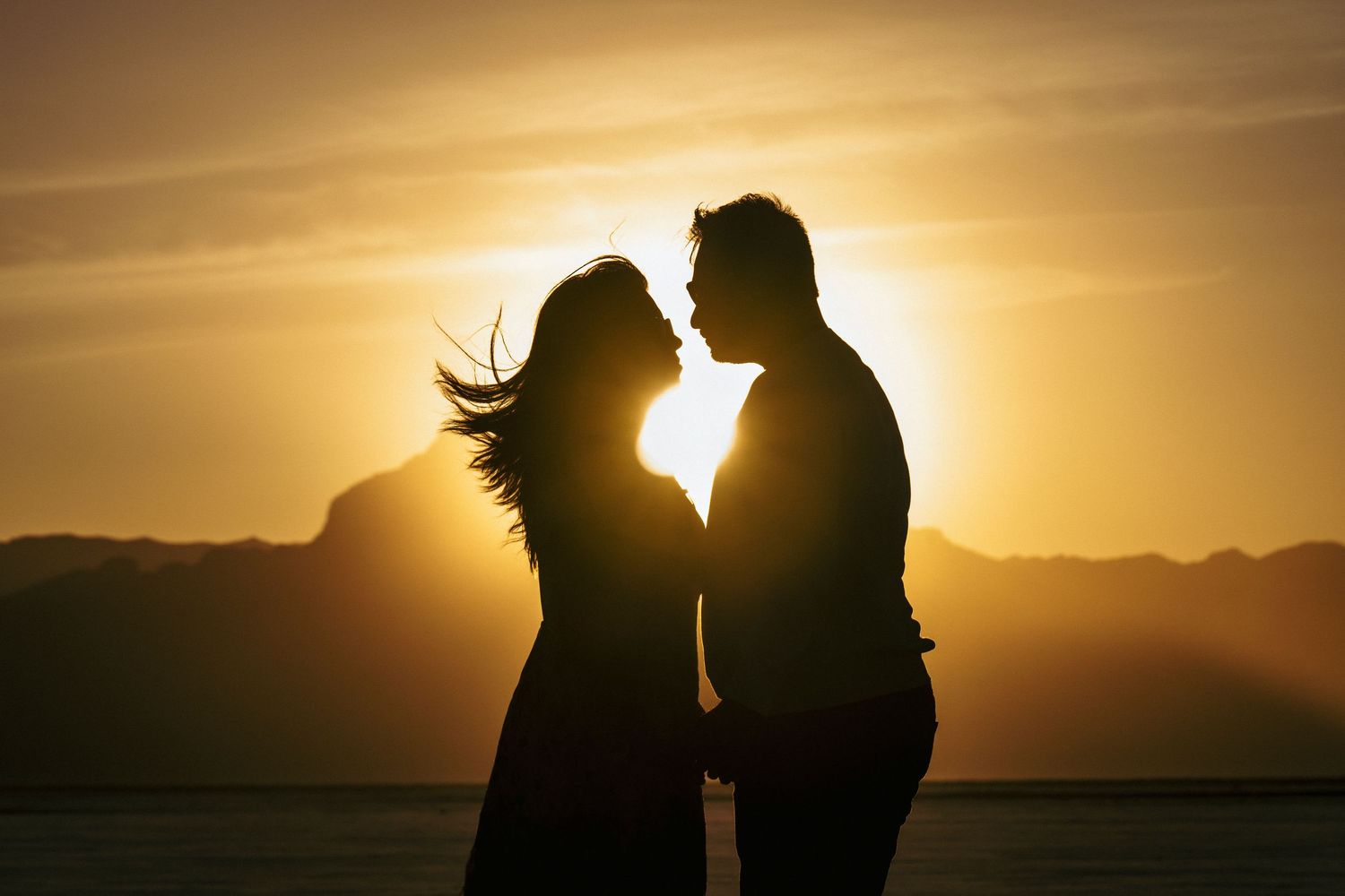 Silhouetted couple standing close together against a dramatic sunset over mountains.