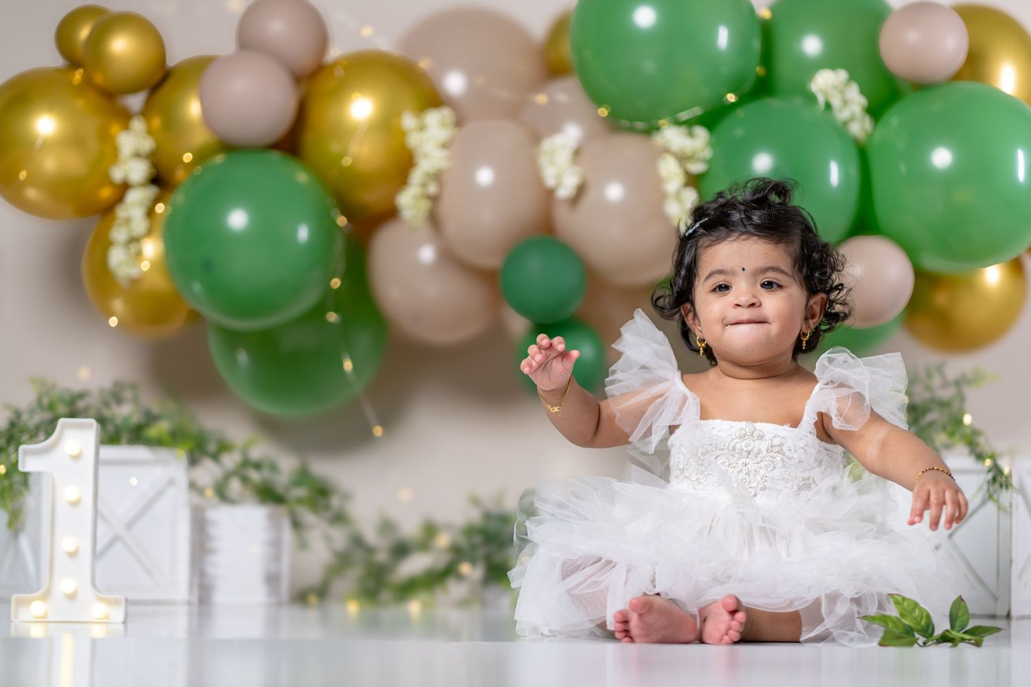 Baby girl in white dress celebrates 1st birthday with green and gold balloon garland backdrop.