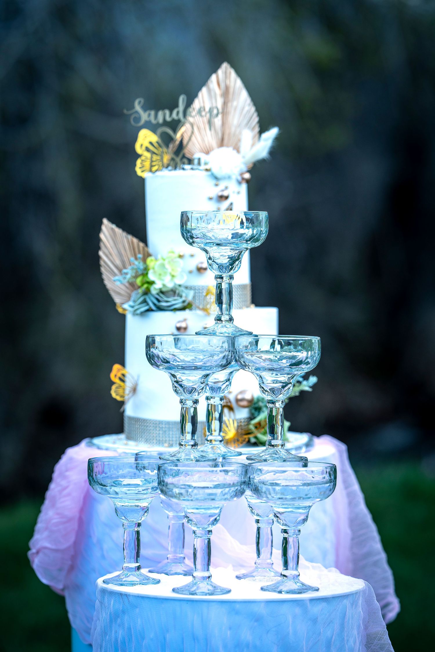 A tiered blue cake with champagne glasses arranged in a fountain display and decorative feather accents.