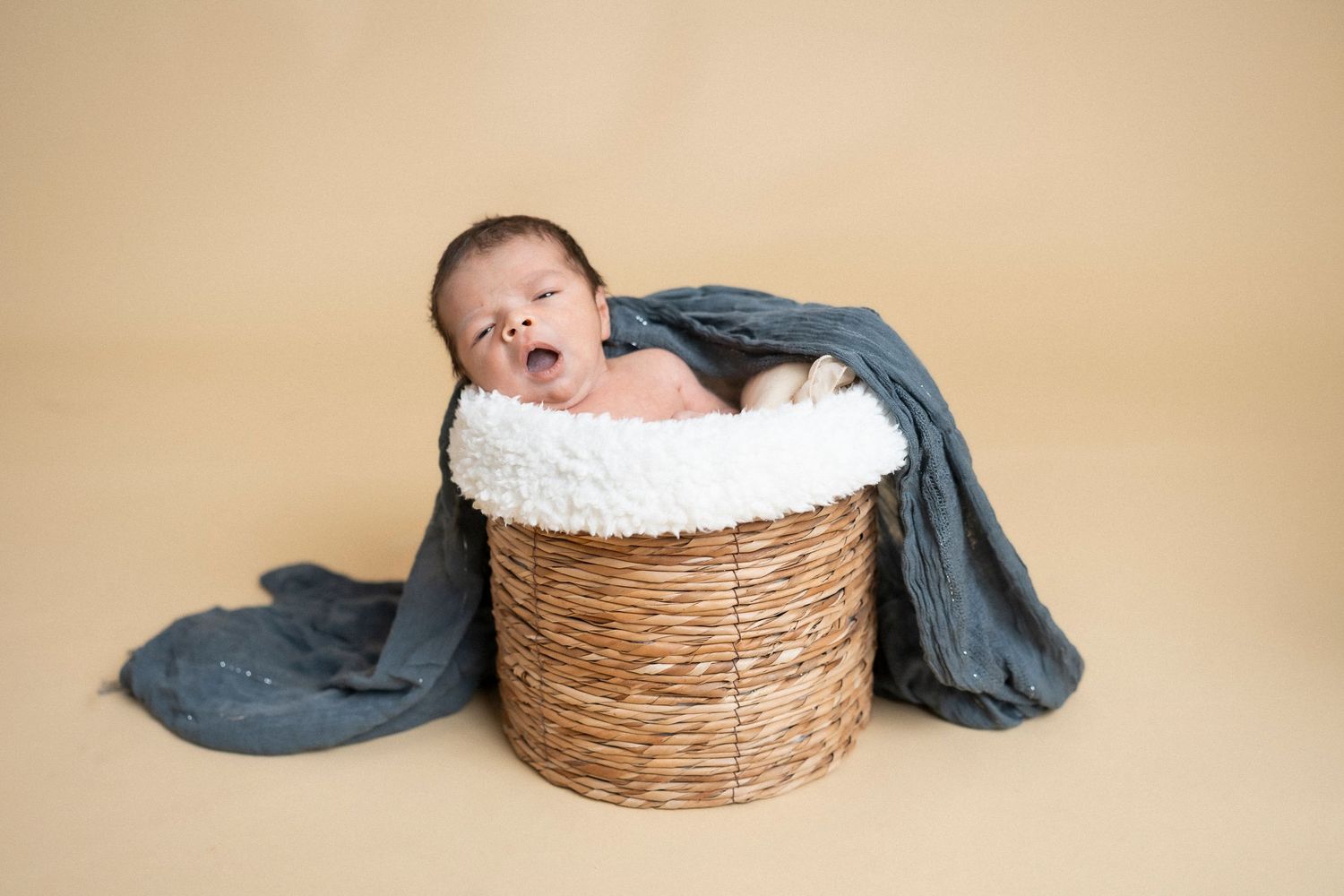 Sleepy newborn baby stretches in cozy woven basket during portrait session.