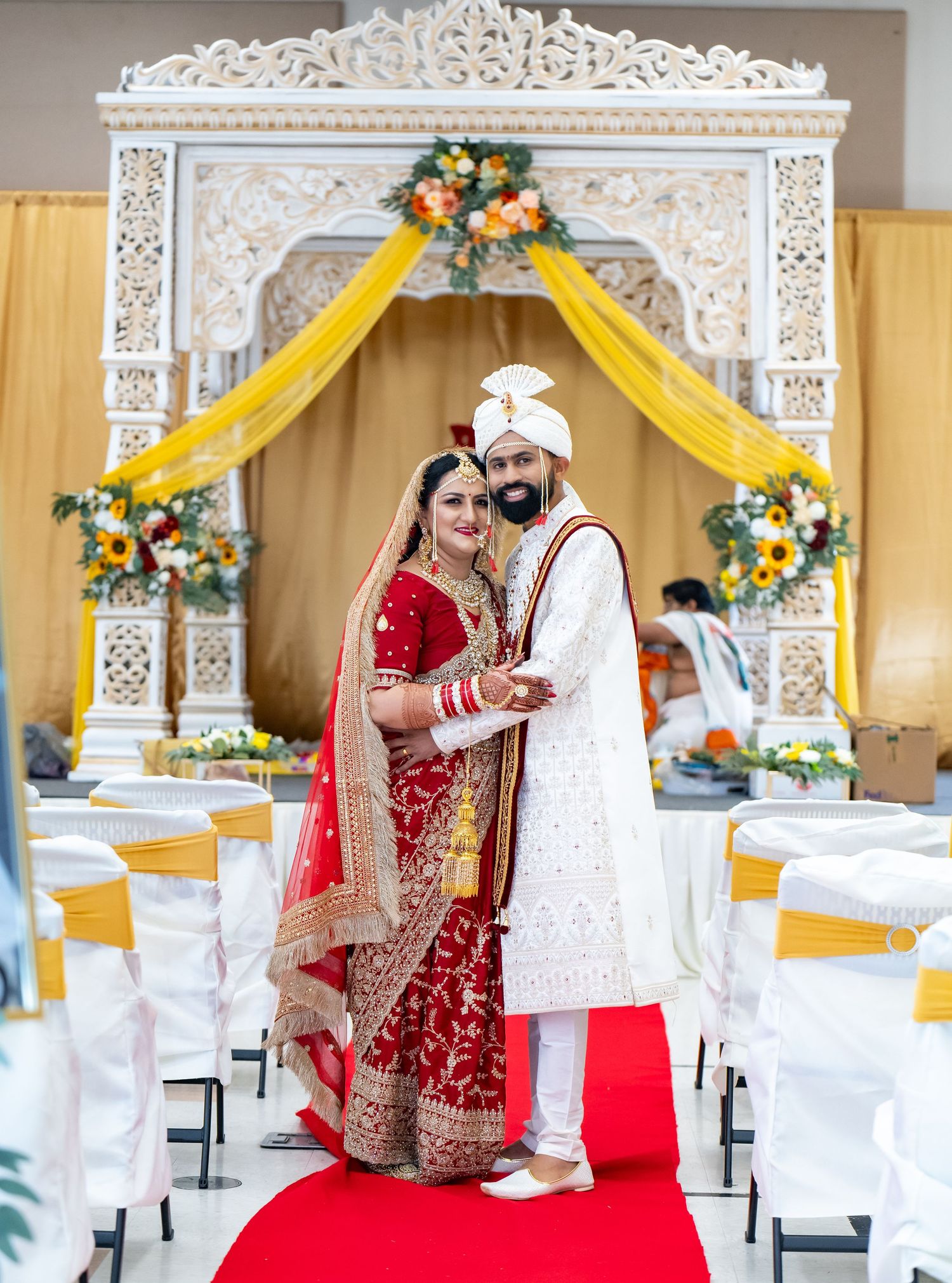 Indian bride in red lehenga and groom in white sherwani pose at traditional Hindu wedding ceremony with floral mandap.