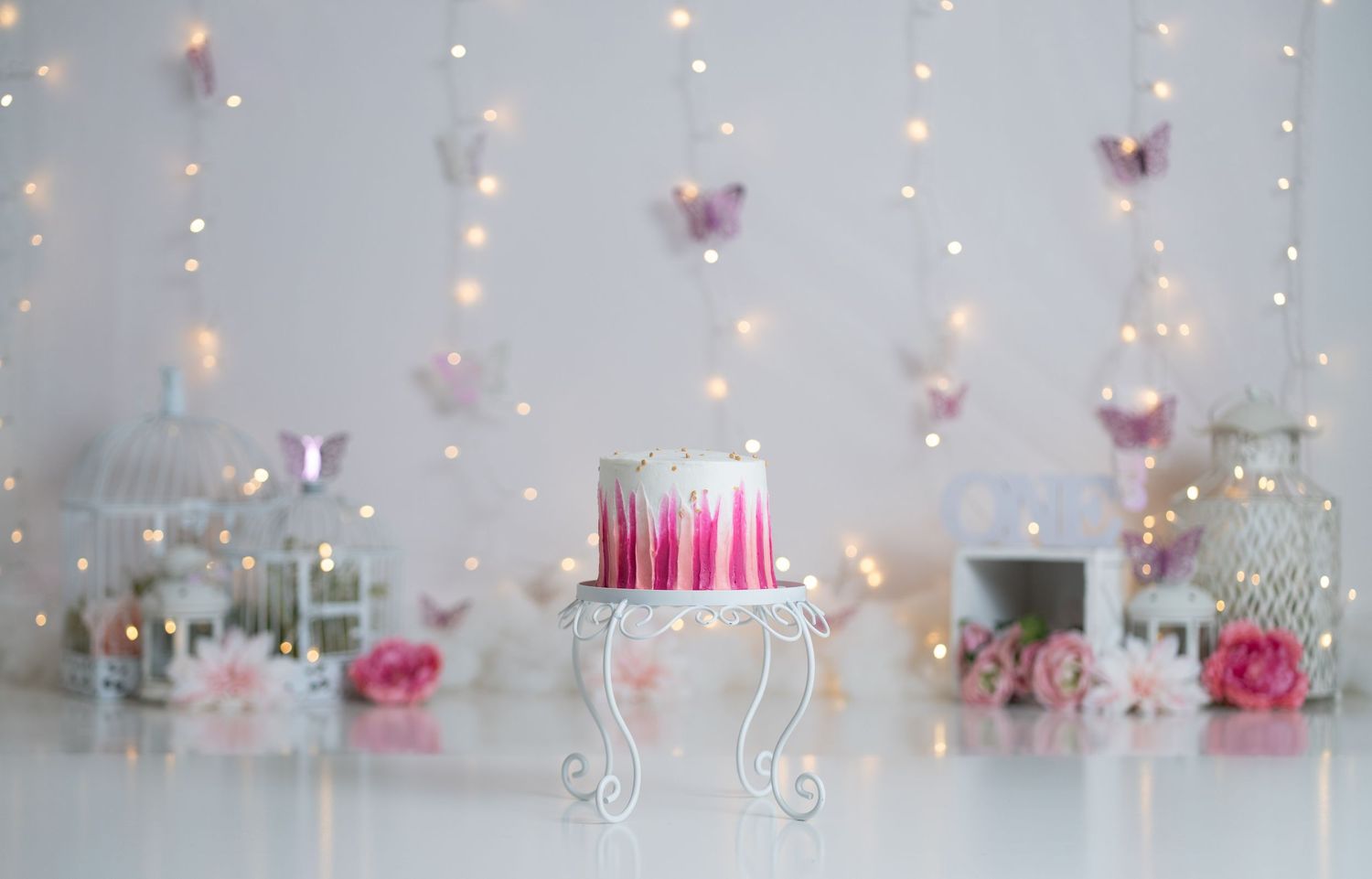 Pink birthday cake on ornate stand with butterfly decorations, fairy lights, and floral accents on white background.