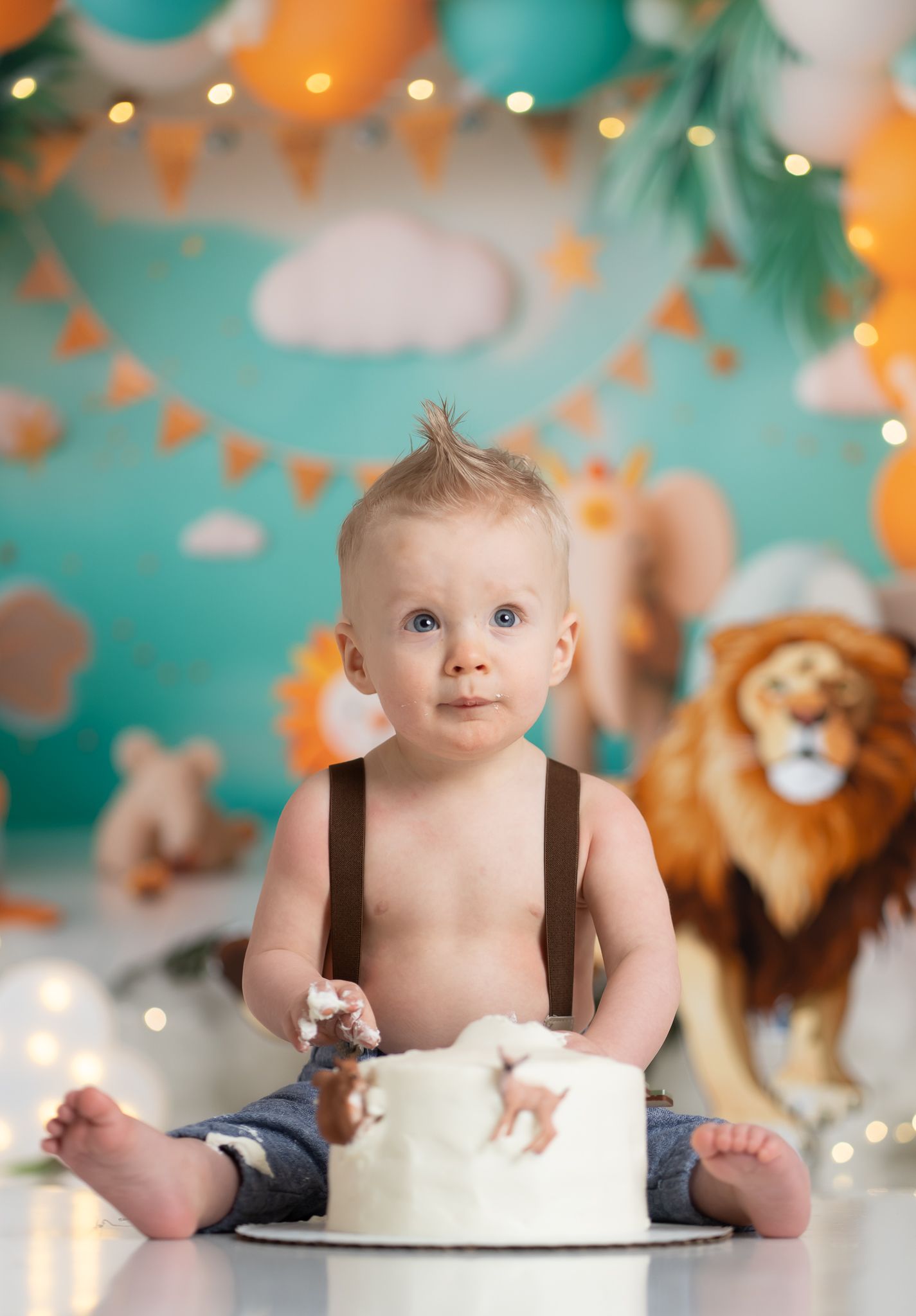 Baby in suspenders with birthday cake against teal backdrop with party decorations.