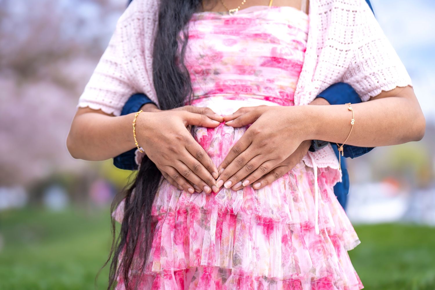 Woman in pink tie-dye dress making heart shape with hands over pregnant belly in garden.