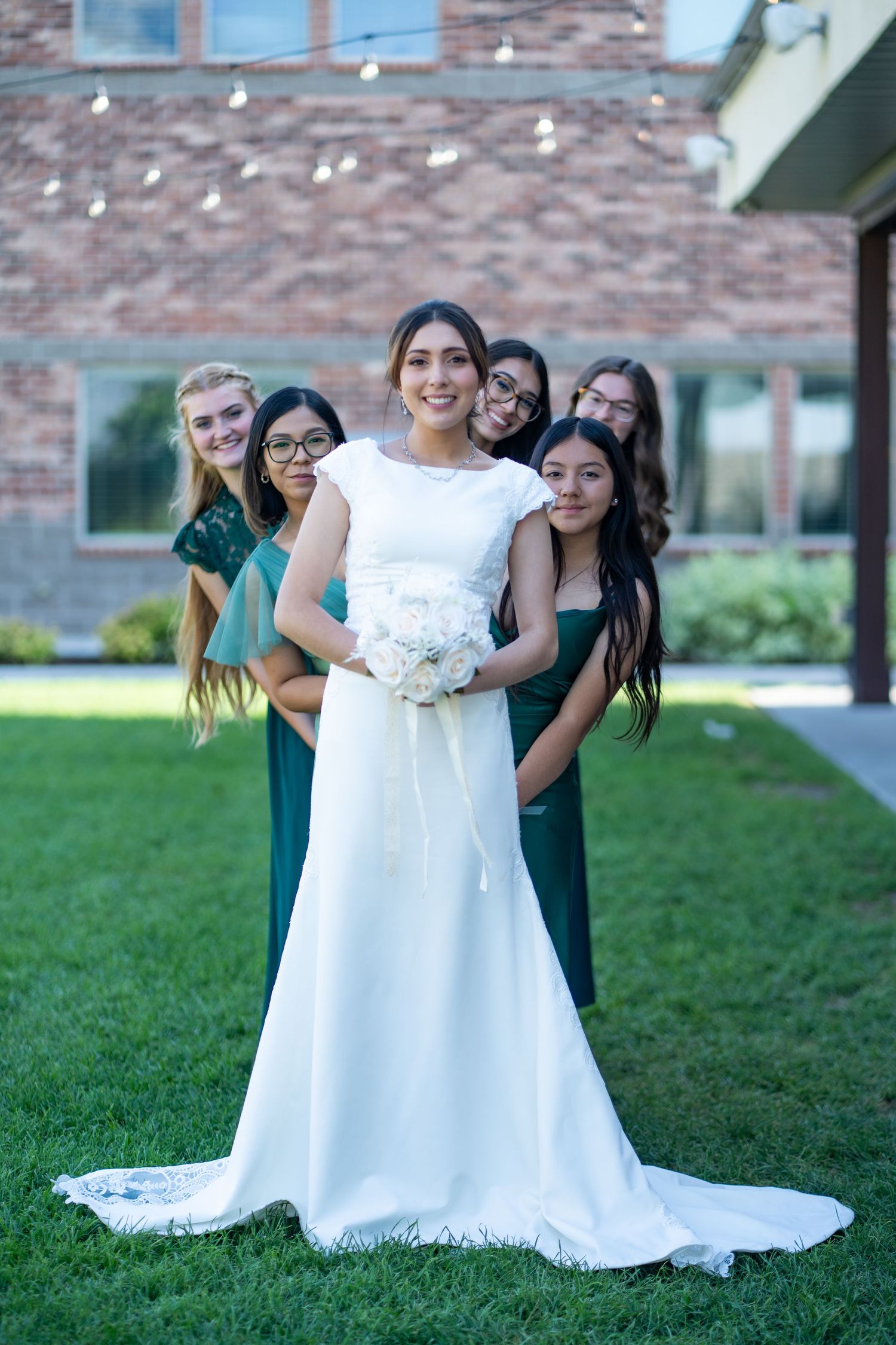 Bridal party in emerald green dresses poses behind bride in elegant white gown outdoors.