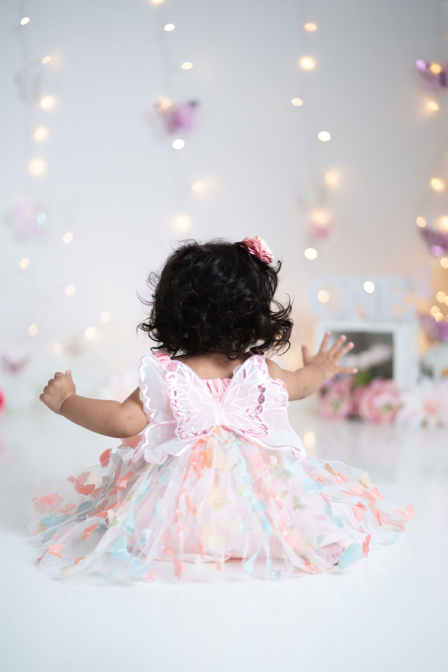 Baby girl in colorful butterfly dress sits on white floor surrounded by bokeh lights and butterfly decorations.