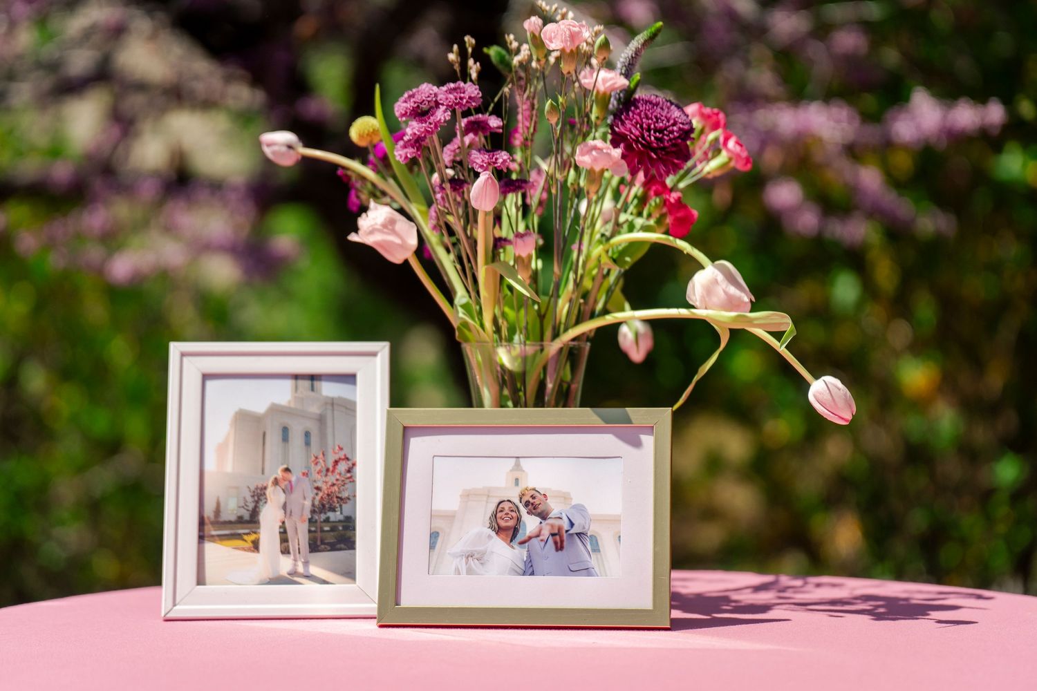 Framed wedding photos displayed with pink and purple floral arrangement on pink table.