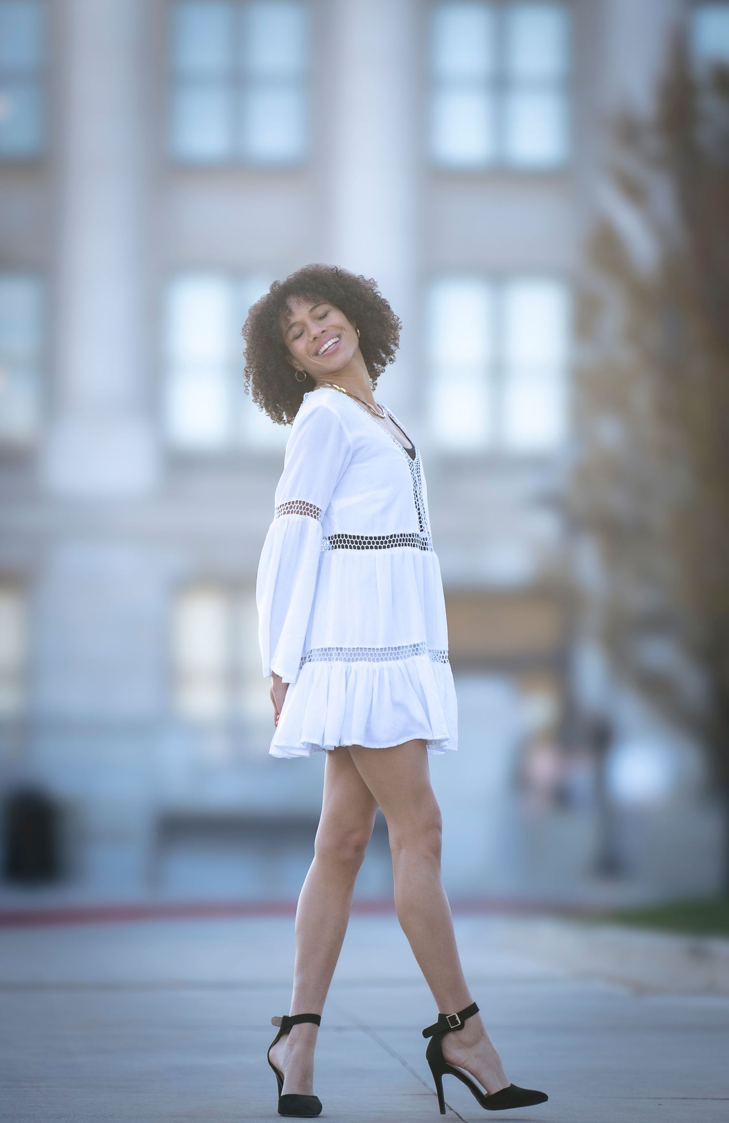 A person in a white flowy dress poses outdoors against a blurred building backdrop.