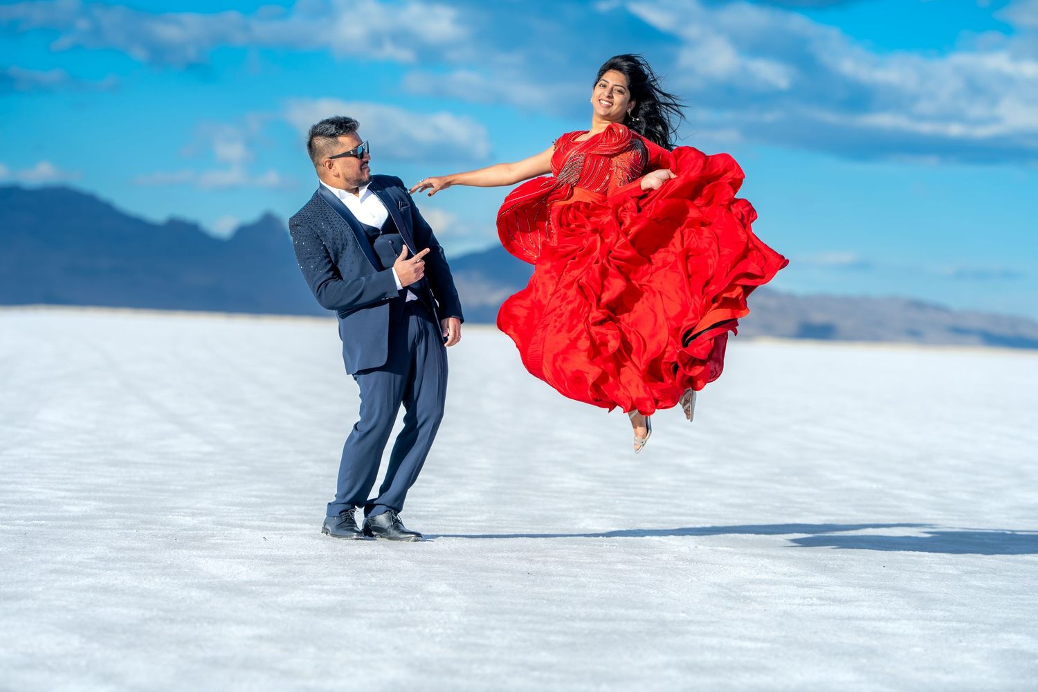 A couple in formal attire poses dramatically on a salt flat with the woman in a flowing red dress leaping in the air.