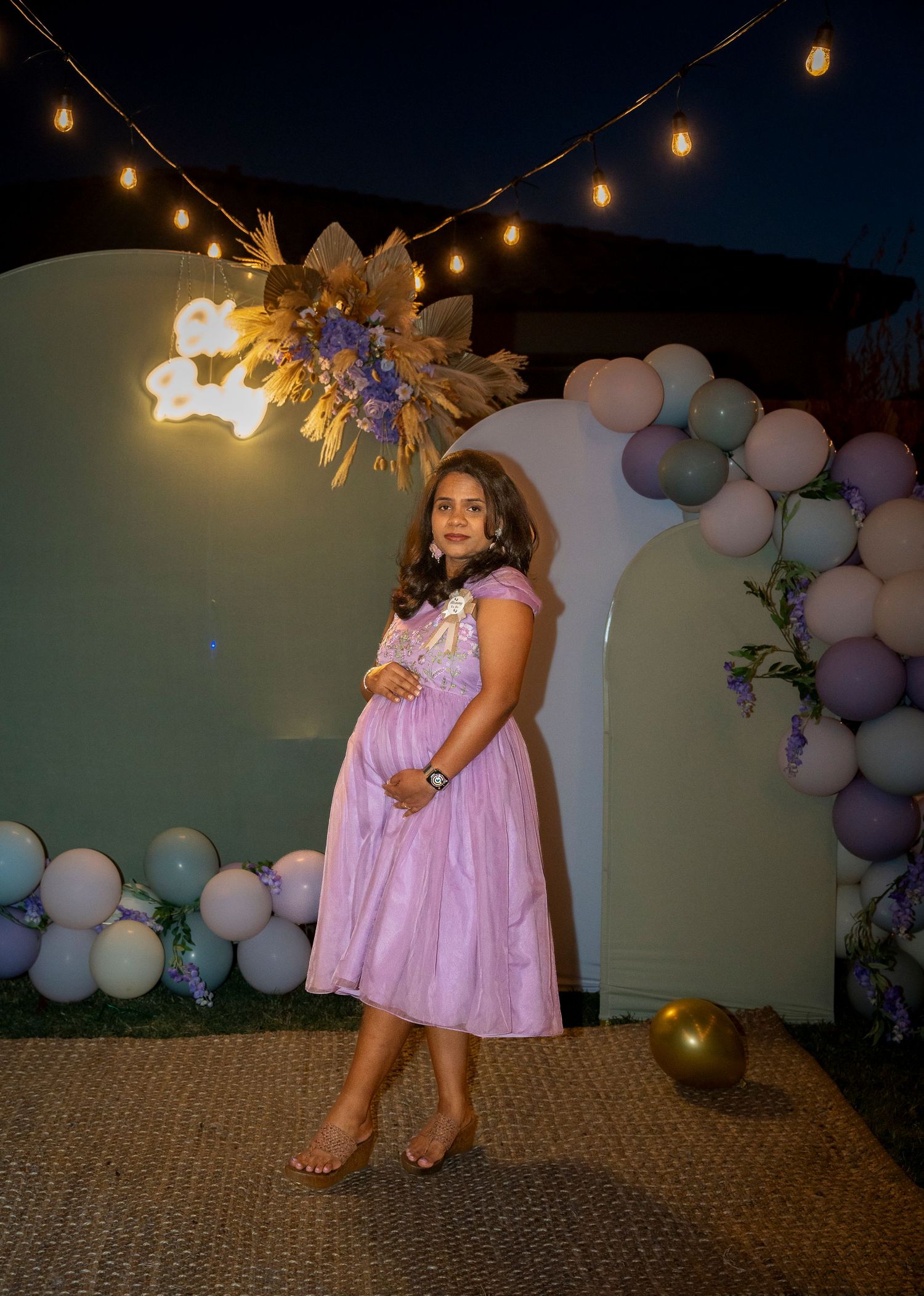 A maternity photo featuring a woman in a lilac dress under string lights and balloon decorations.