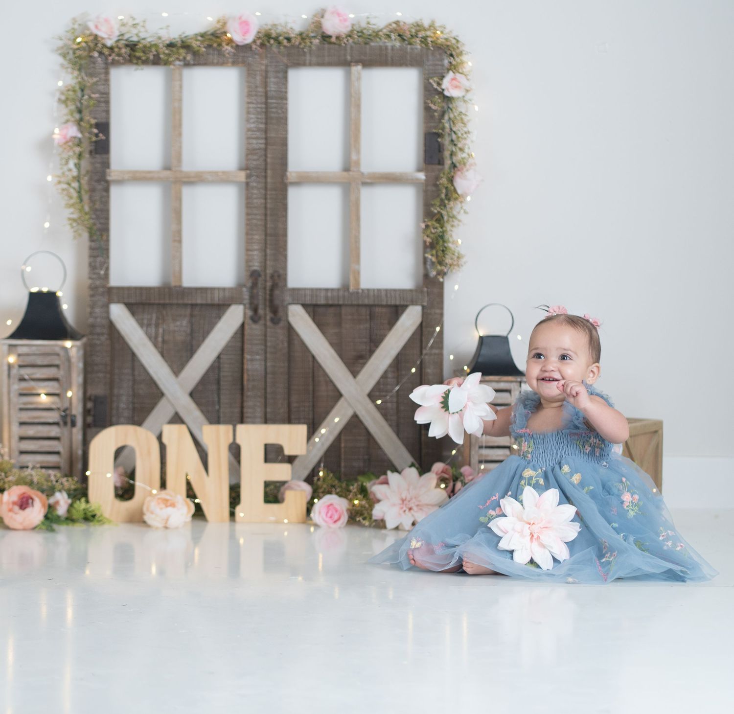 Baby girl in blue dress sits beside ONE letters at rustic barn door first birthday cake smash photo session.