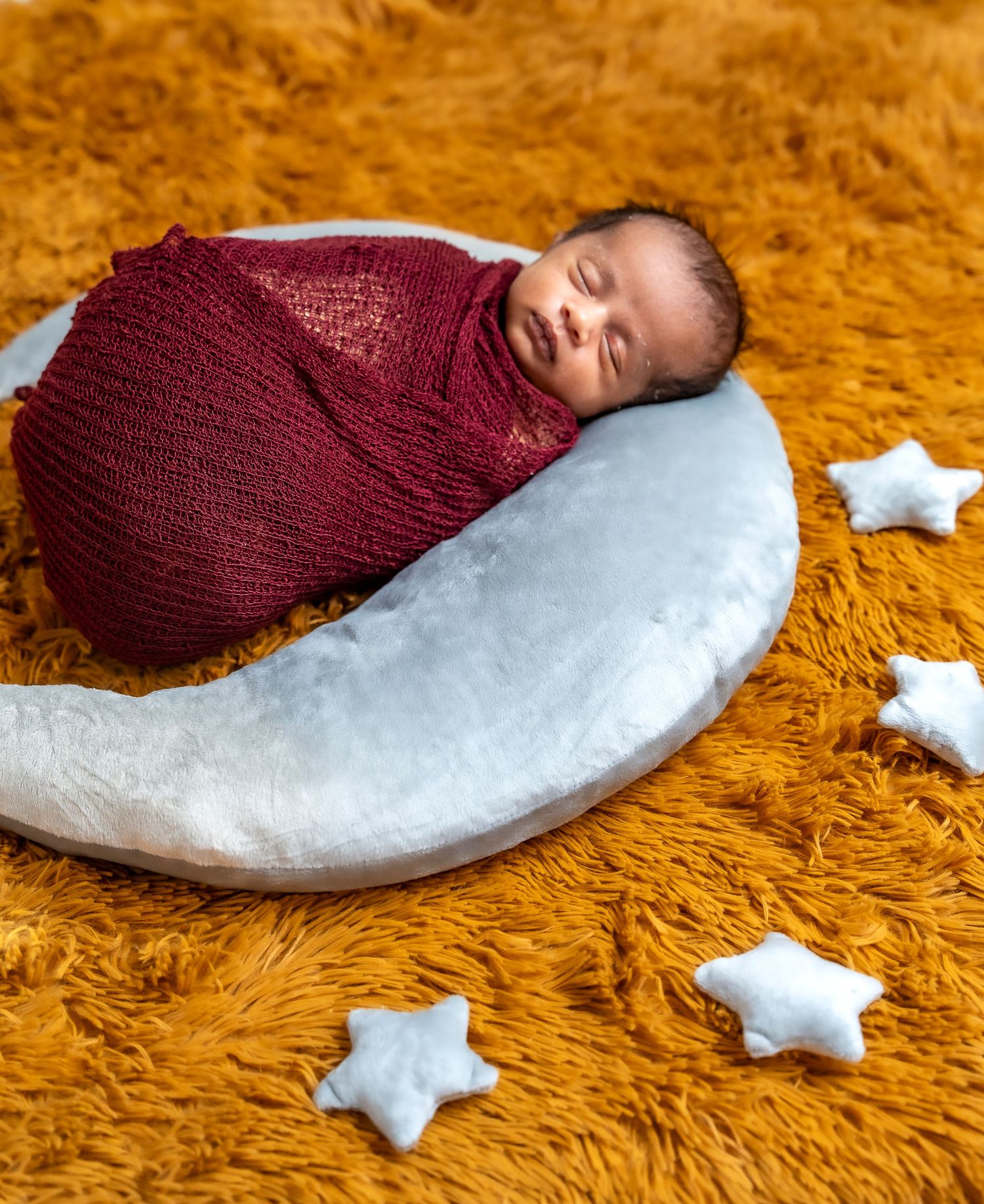 A sleeping newborn wrapped in burgundy knit swaddle rests on a crescent moon prop with white stars on orange carpet.