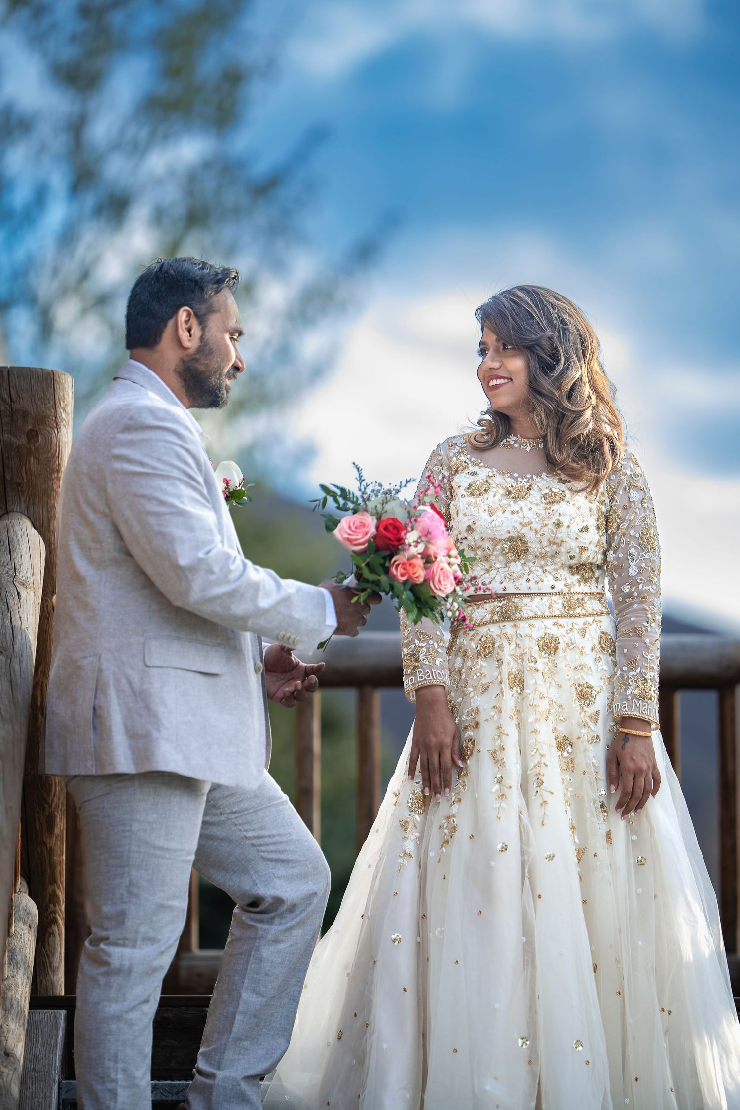 Couple in wedding attire share a romantic moment on a rustic outdoor deck with the bride holding pink flowers.