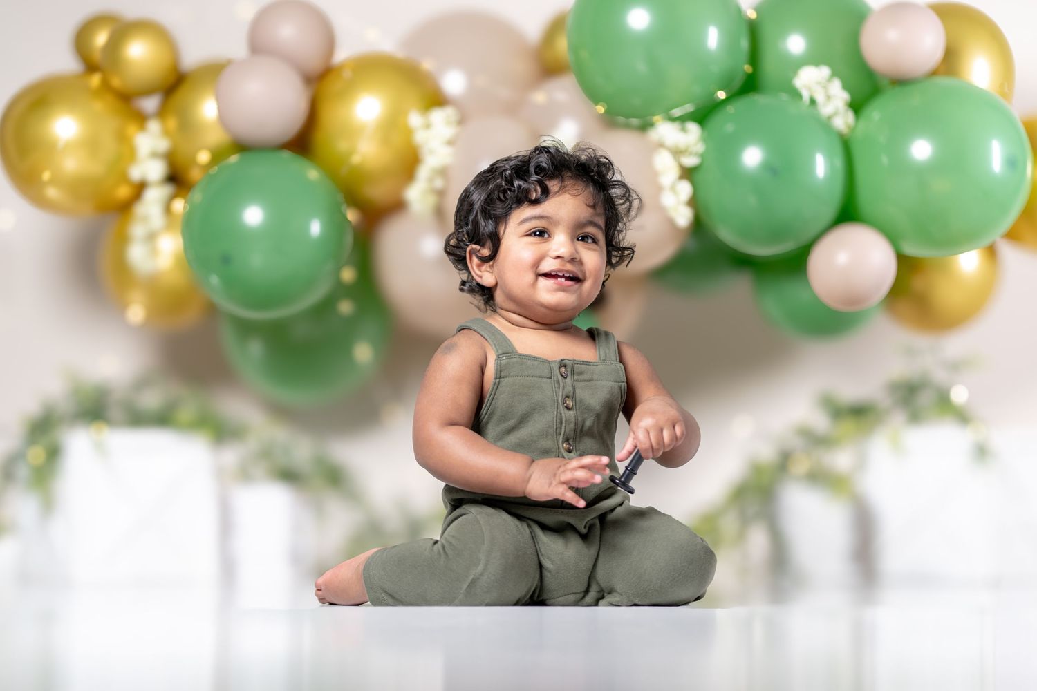 Happy toddler in green outfit sitting before balloon backdrop with gold, green, and beige balloons at first birthday celebration.