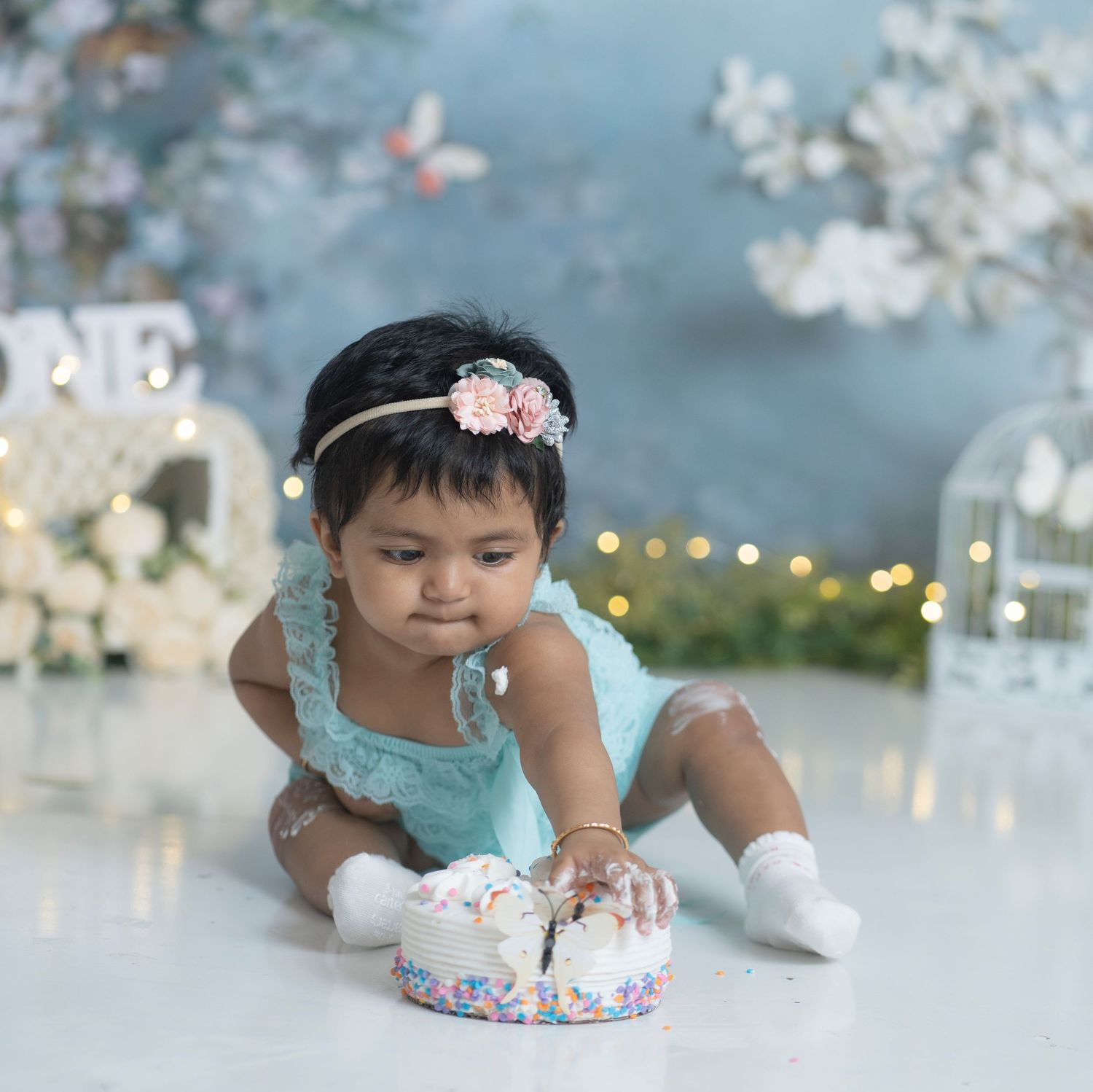 First birthday cake smash photo session with decorative lights and blossoms in background.