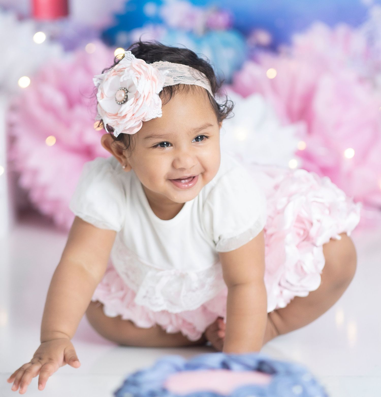 Baby photoshoot with white dress, floral headband, and pink tulle against sparkly background.