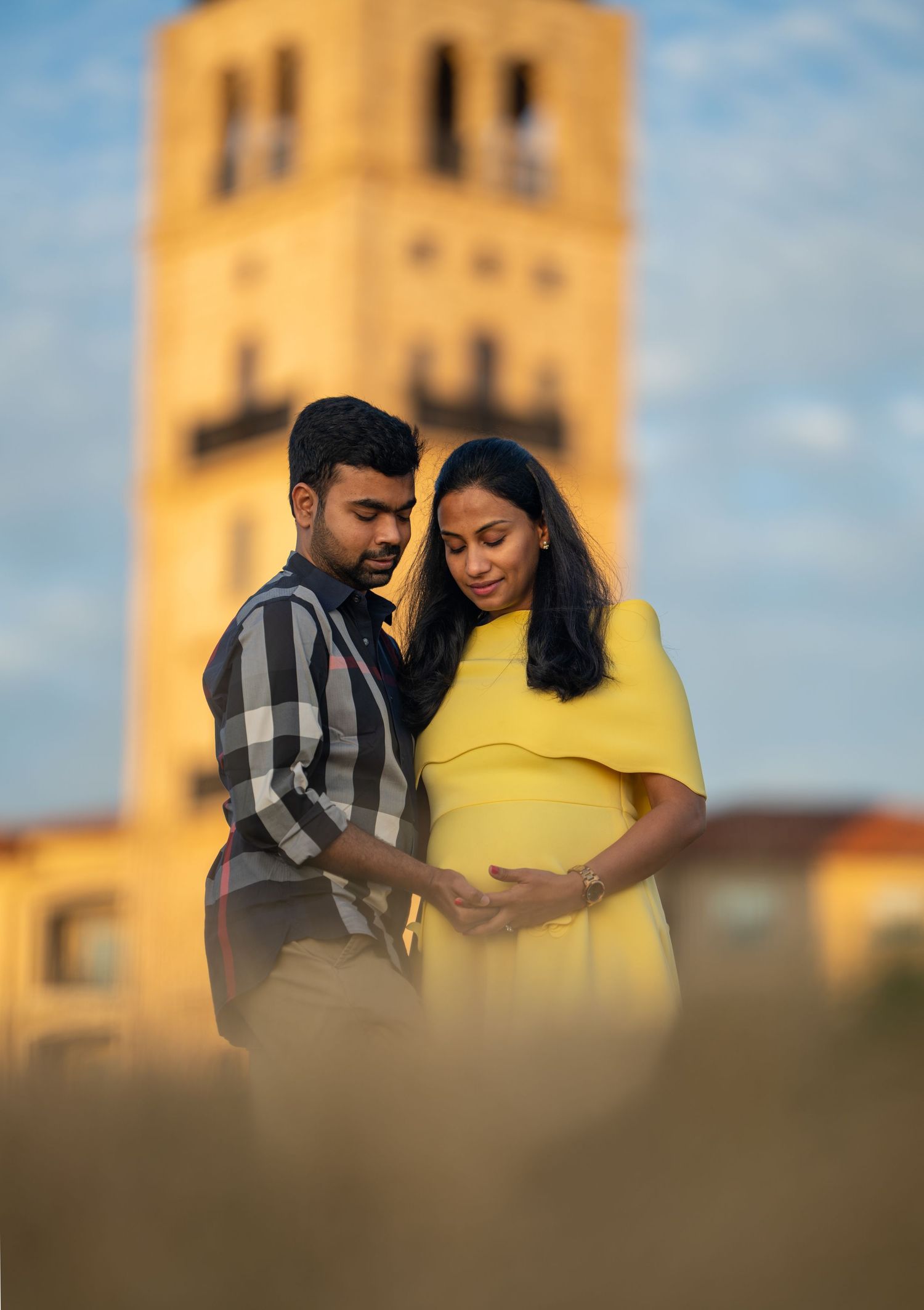 Couple tenderly cradling pregnant belly outdoors at golden hour with blurred tower landmark in background.