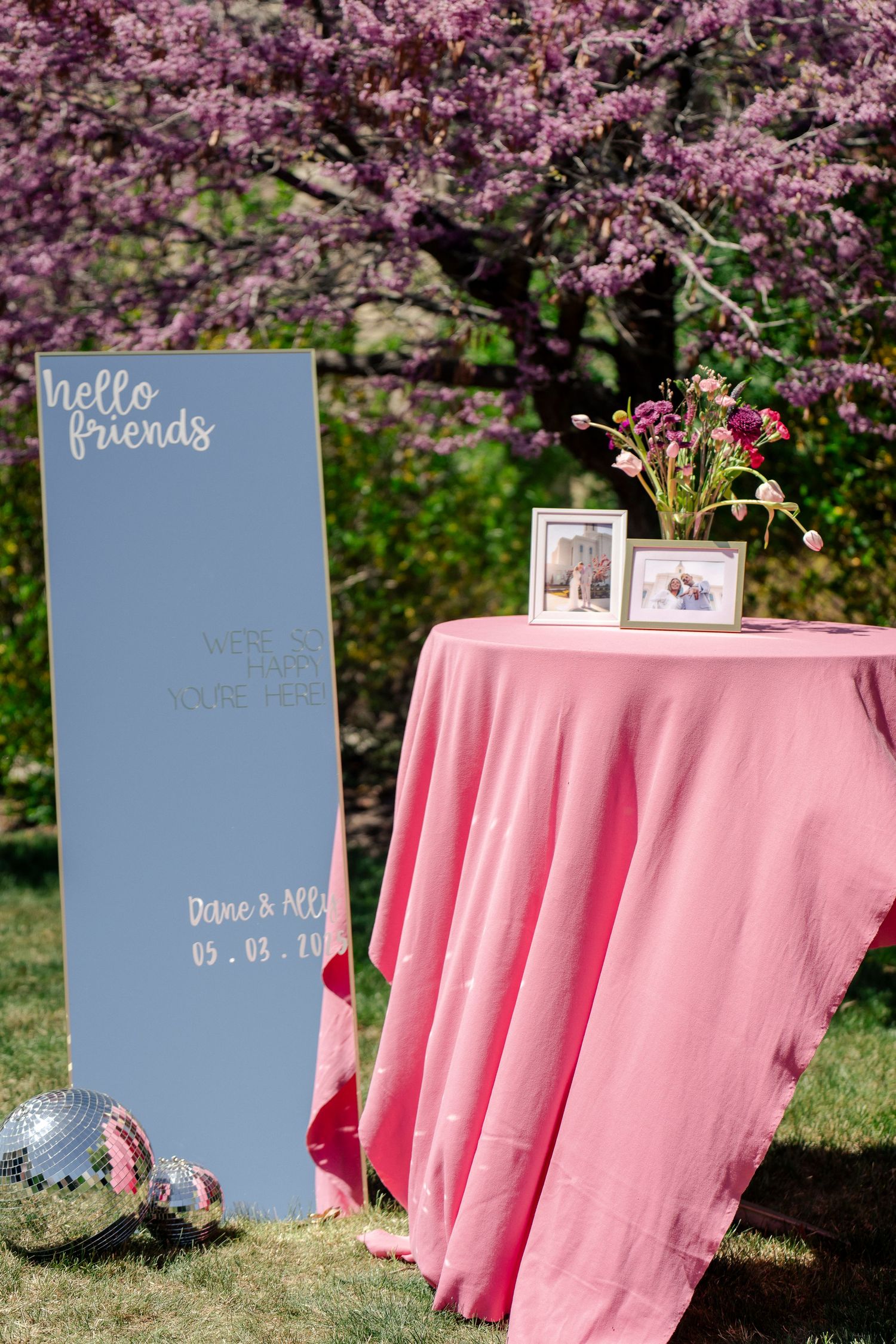 A welcome sign and pink-clothed table with photo frames set against blooming cherry trees.