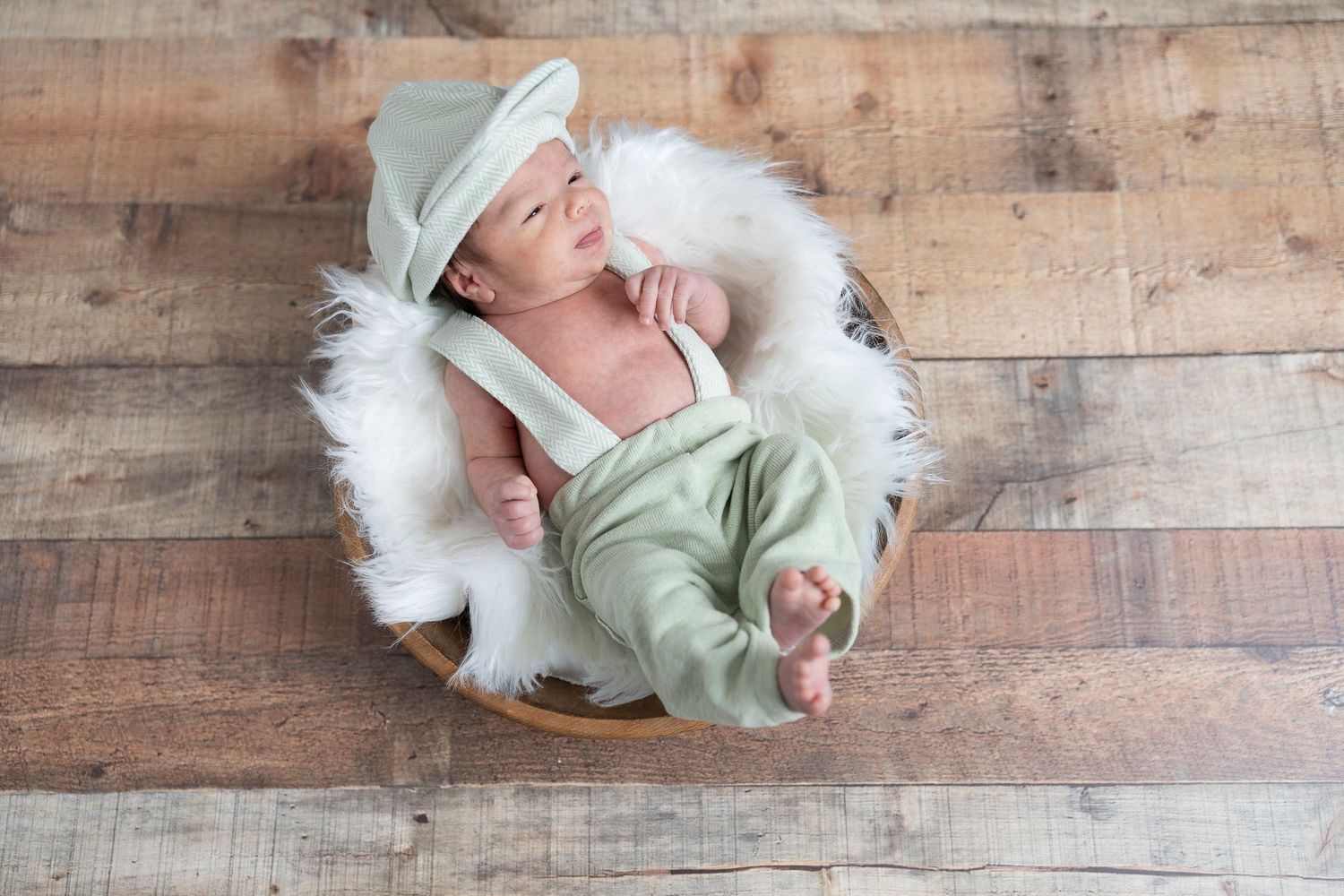 Sleeping newborn wearing mint bonnet on round wooden platform with white fur.