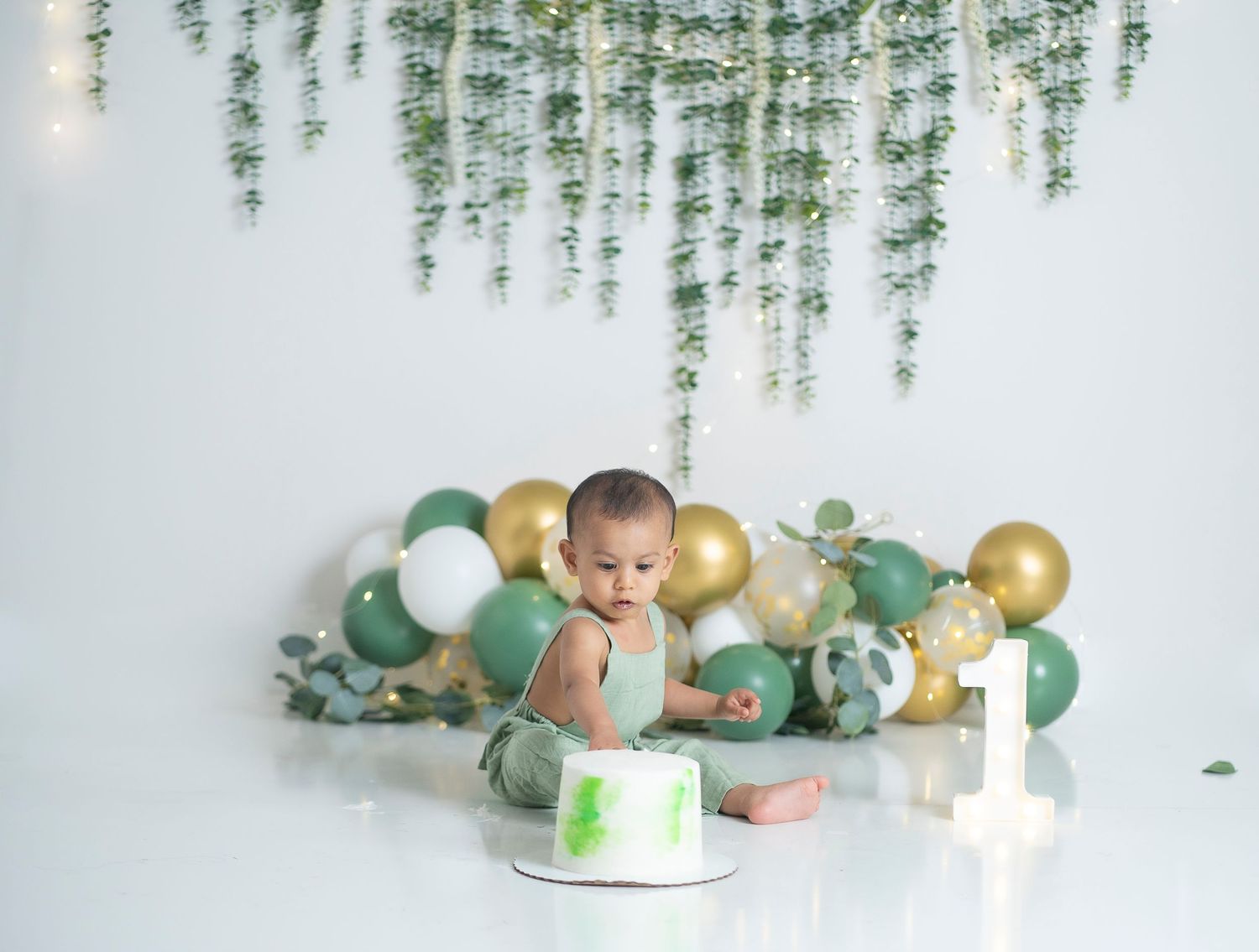 First birthday celebration setup with green and gold balloons, eucalyptus garland, and smash cake on white backdrop.
