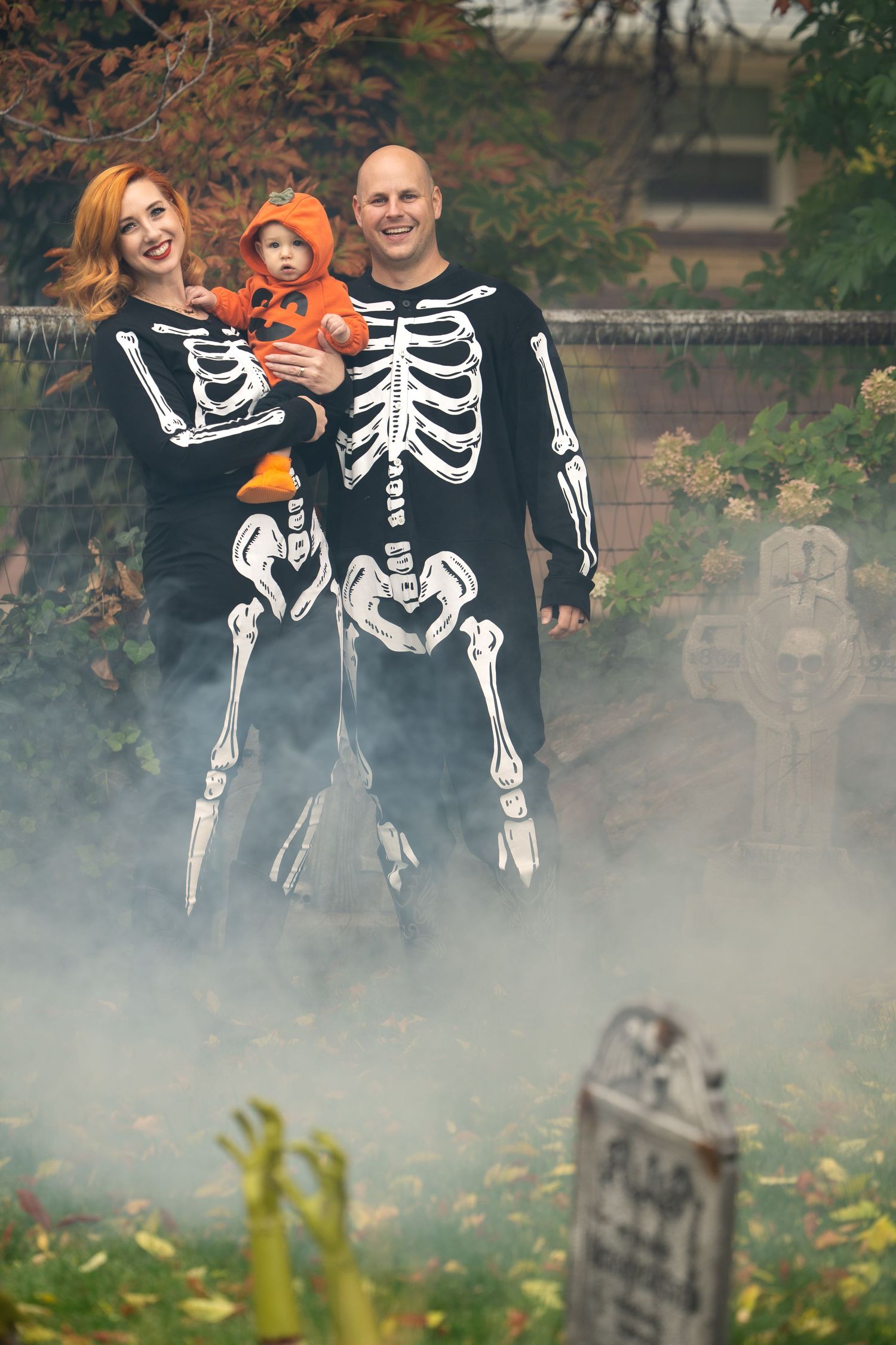 Family wearing skeleton costumes poses together in foggy graveyard scene for Halloween photo.