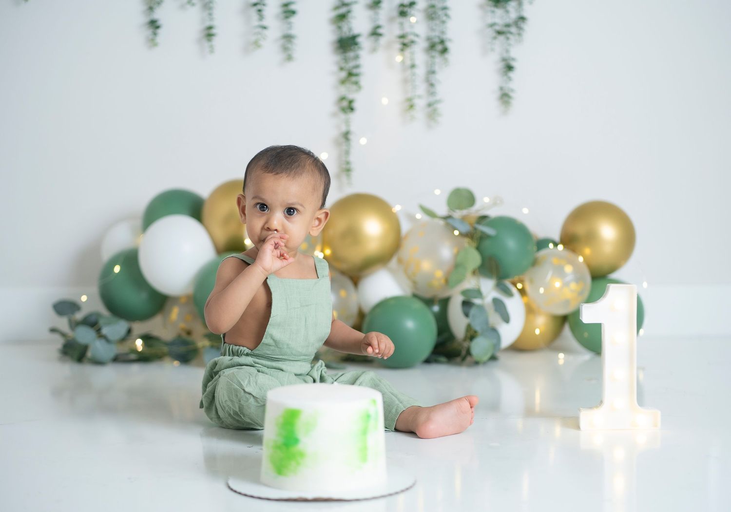 Baby in mint green outfit celebrates first birthday with cake and gold and green balloon decor.