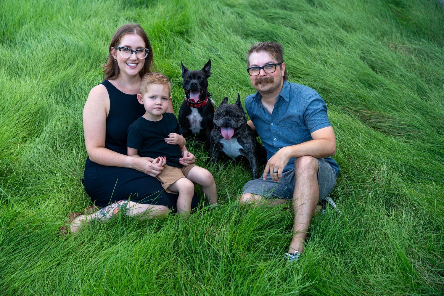 Family portrait with two dogs sitting in tall green grass on a sunny day.