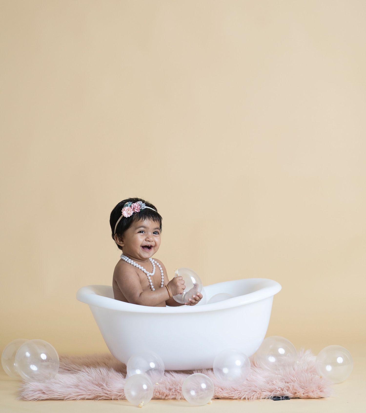 Happy baby sitting in white bathtub with floating bubbles and pink rug.