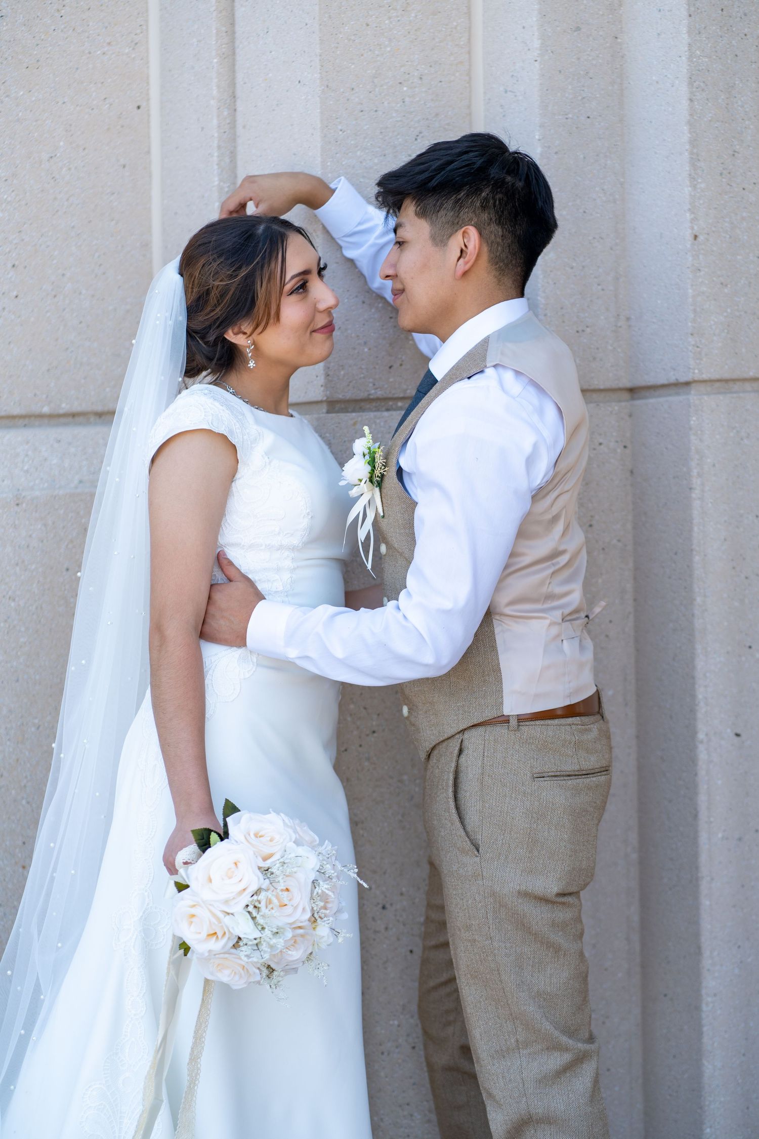 Romantic wedding couple in white and beige outfits sharing intimate moments against a stone wall.