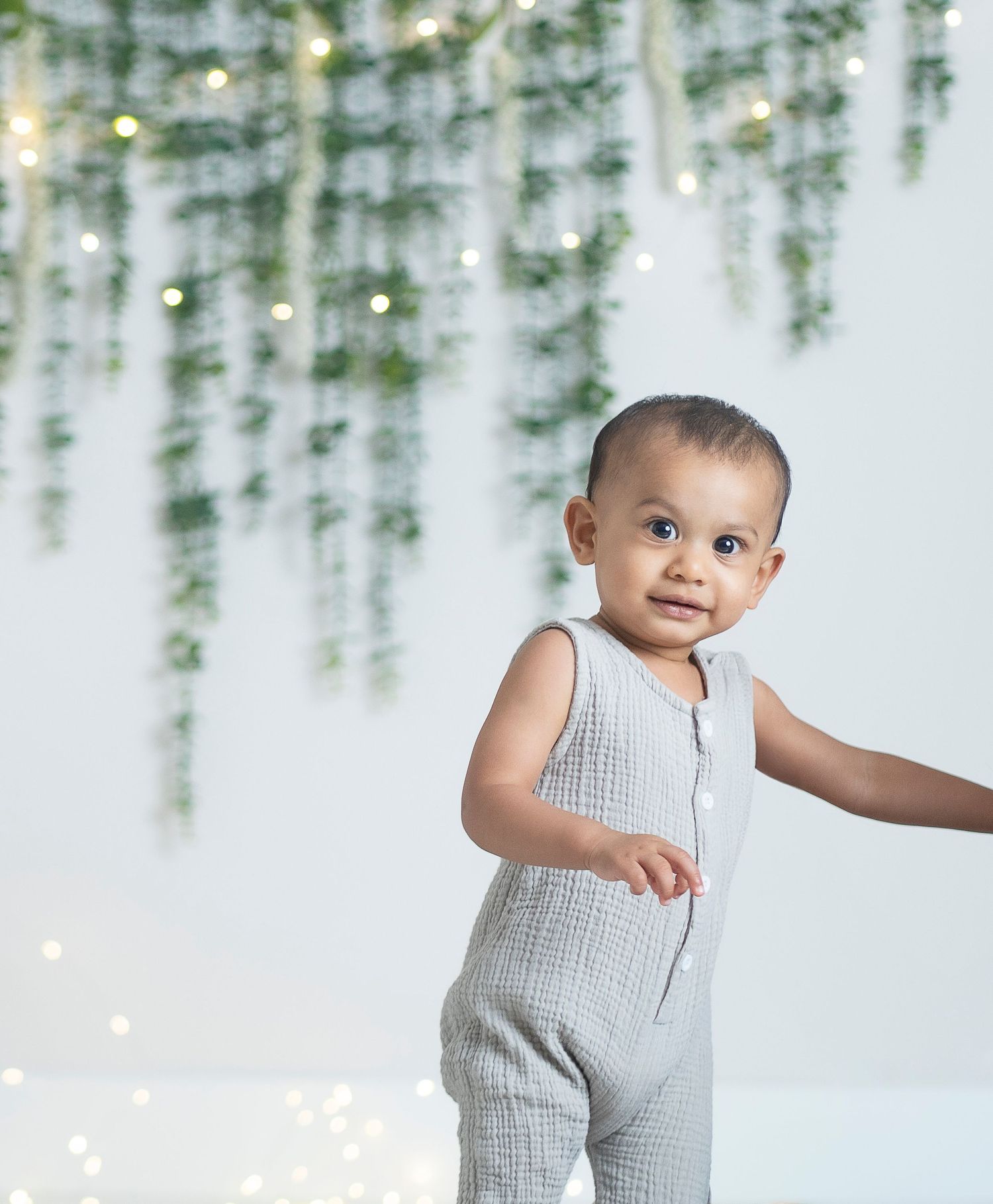 Baby in grey romper standing against white backdrop with hanging greenery and lights.
