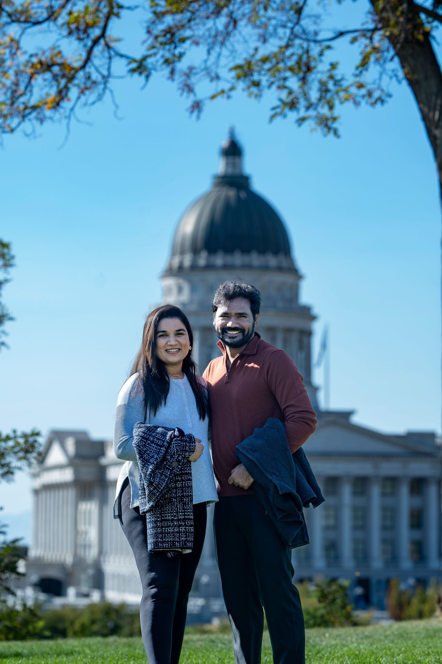 Couple standing on grass lawn with Utah State Capitol building dome in background.