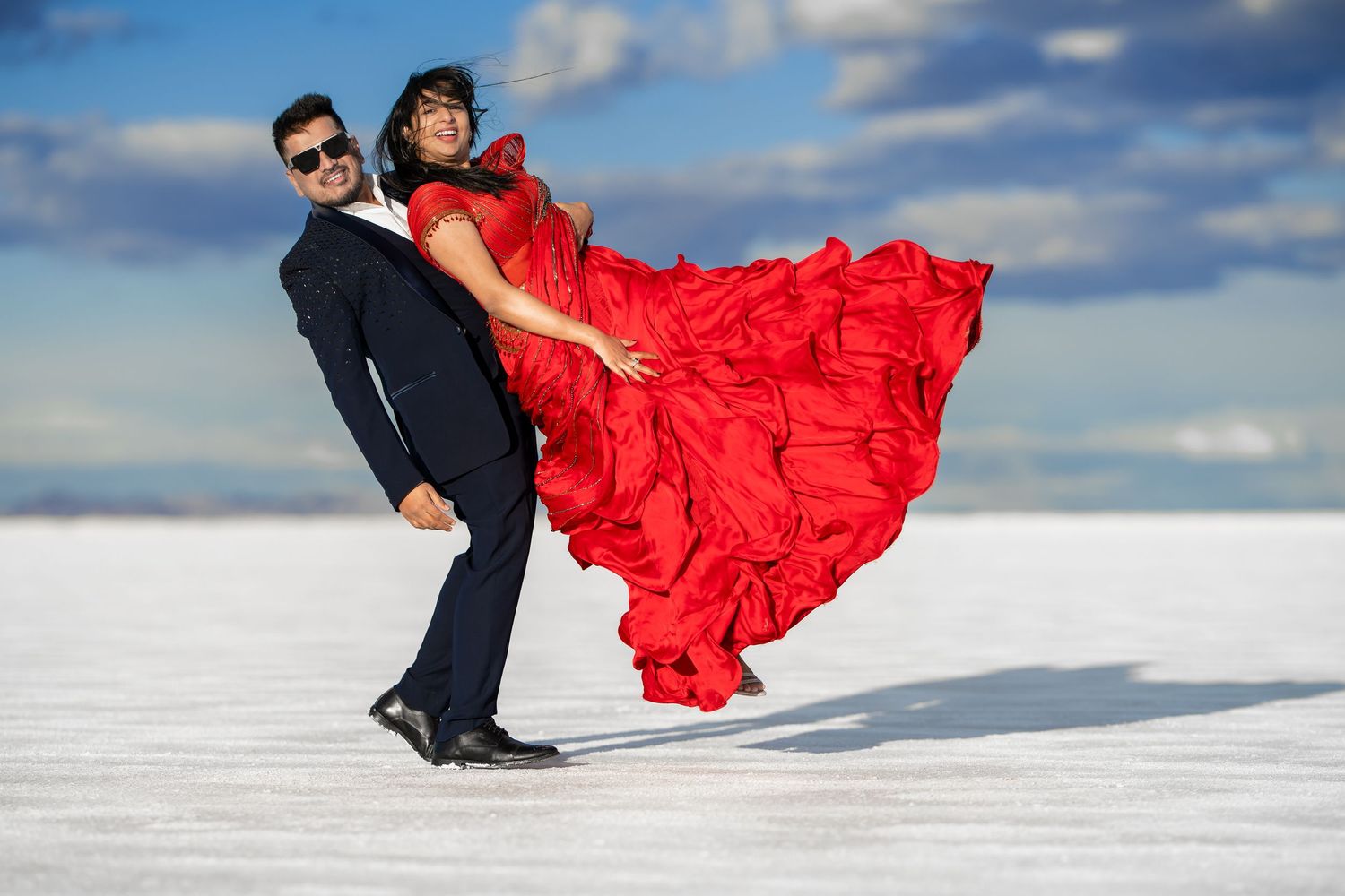 Red dress flows dramatically as couple poses on salt flats under blue sky.