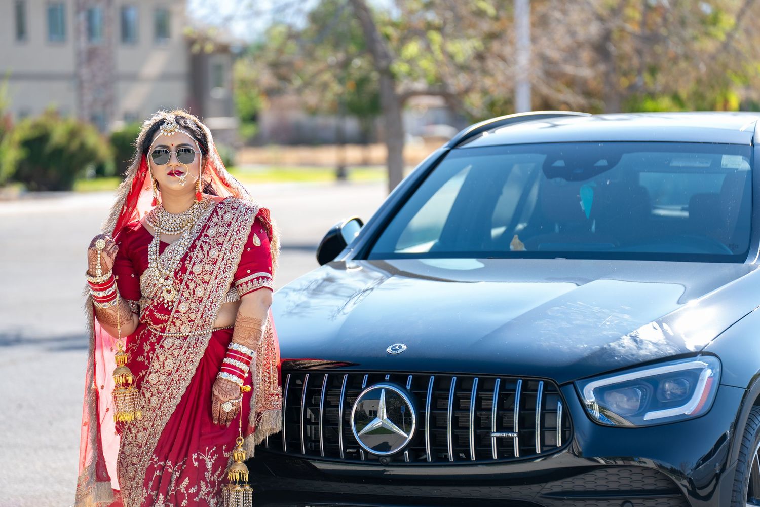 Indian bride in red bridal saree and jewelry poses beside a Mercedes-Benz SUV outdoors on a sunny day.