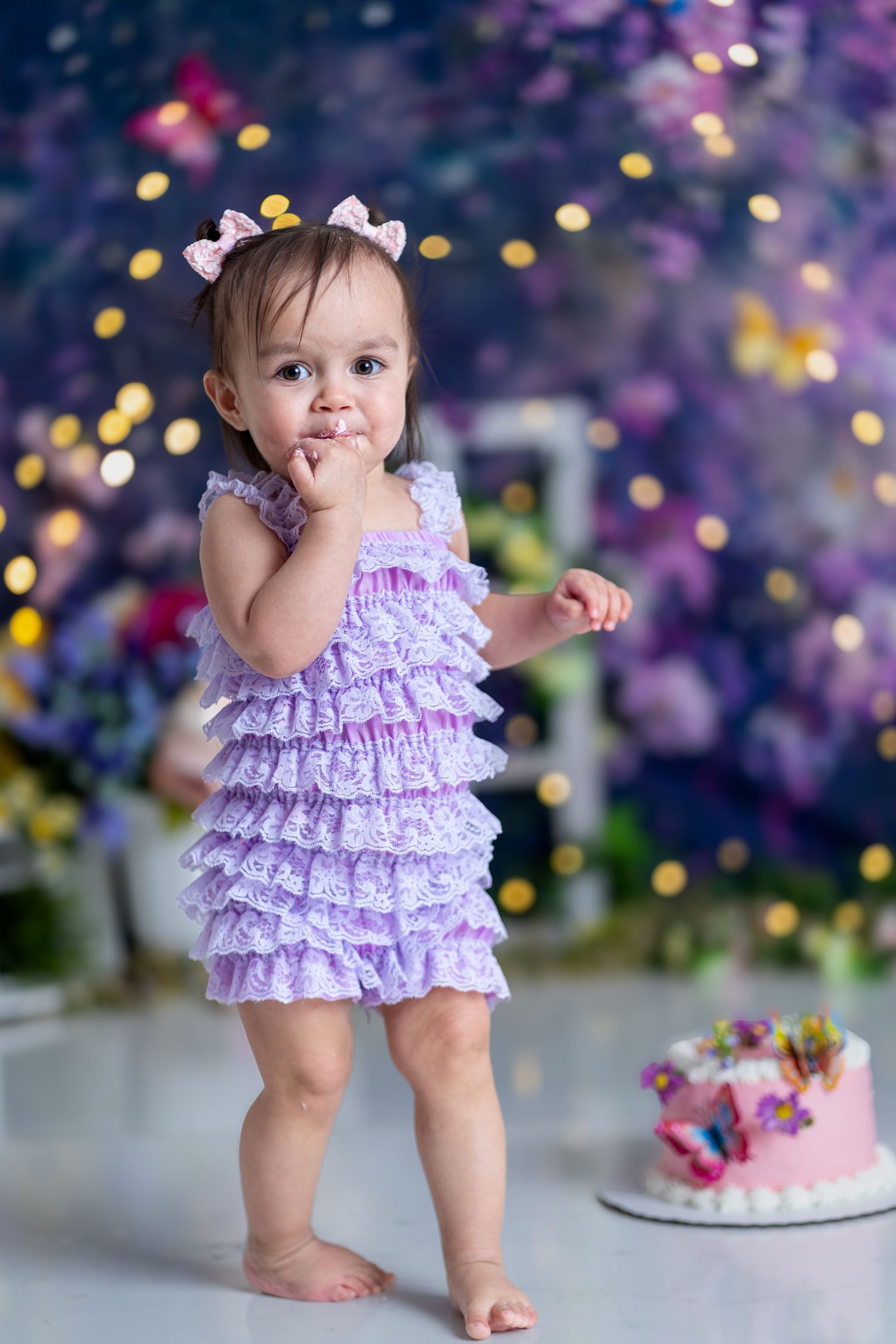 Toddler in purple ruffle dress standing by birthday cake with bokeh lights.
