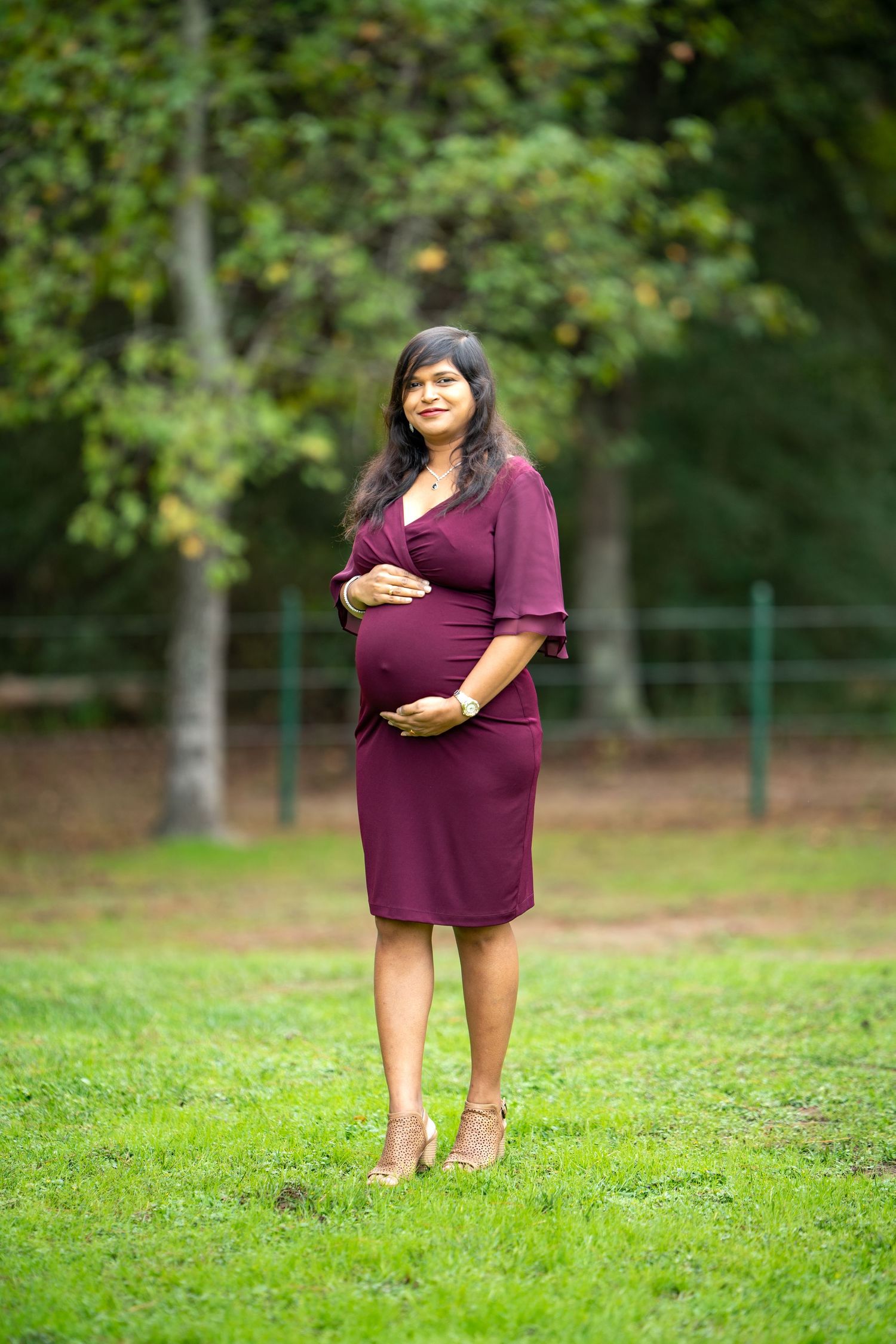 A pregnant woman in a burgundy dress stands on grass in a park setting.