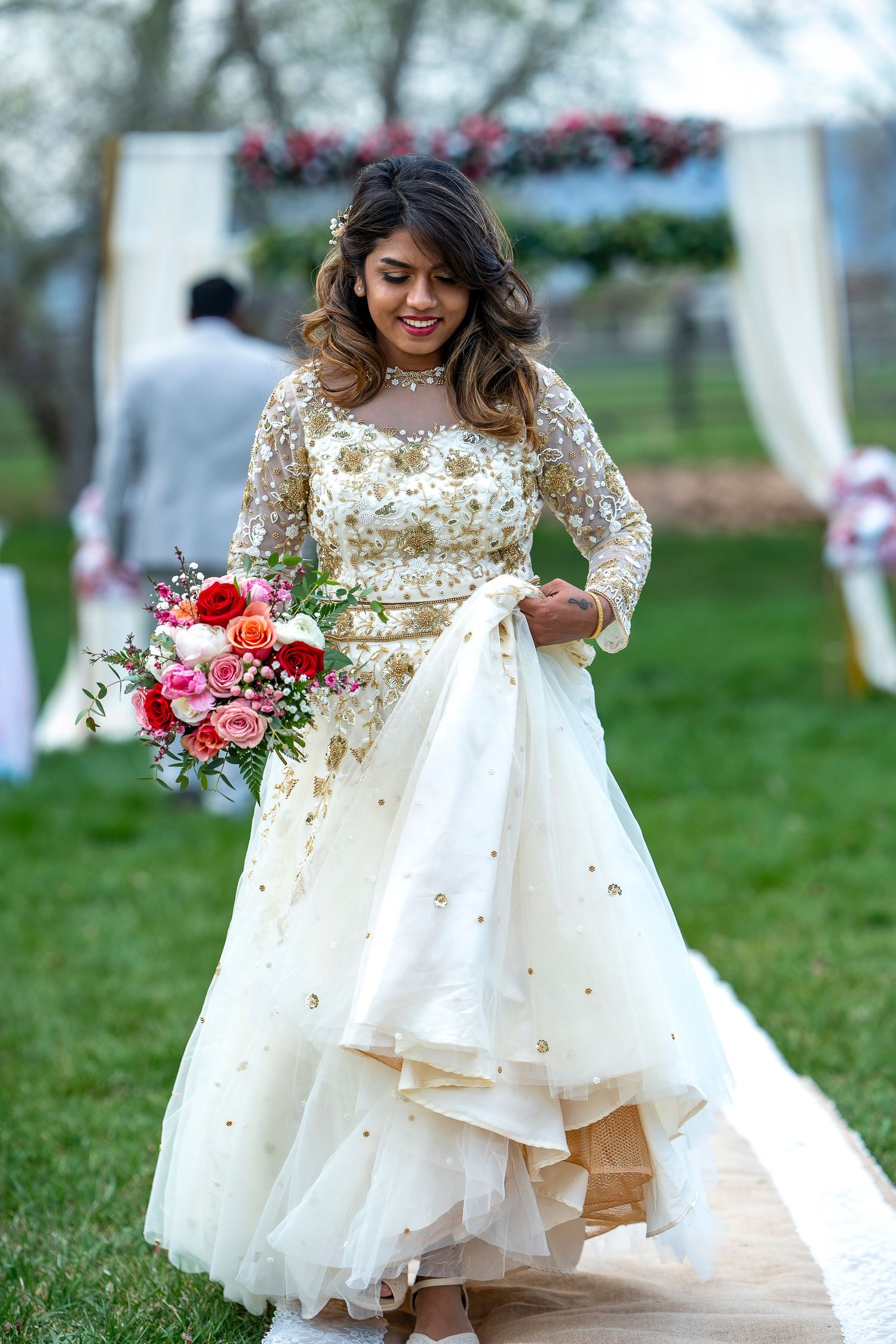 Bride in embellished white gown holding colorful bouquet at outdoor ceremony.
