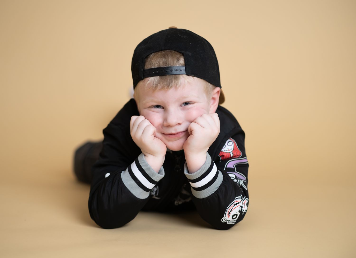 A child wearing a black baseball cap and striped sleeves lies on a beige background in a playful pose.