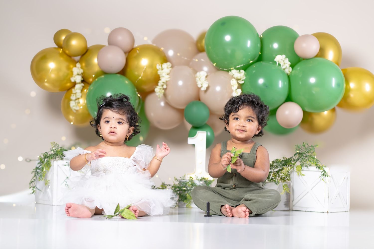 Two toddlers celebrate their first birthday with green and gold balloon backdrop, wearing white dress and green outfit.