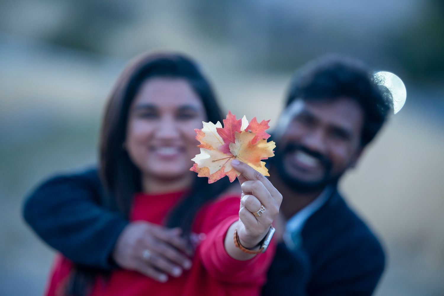 Couple holding autumn maple leaf with blurred background in romantic outdoor setting.
