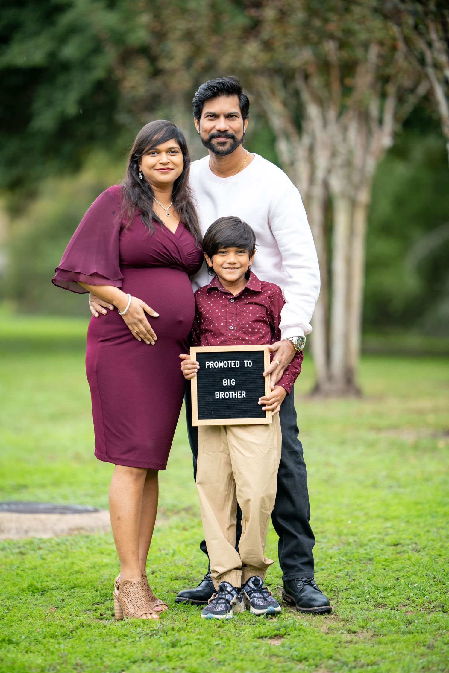 A family poses outdoors with a young boy holding a pregnancy announcement sign.