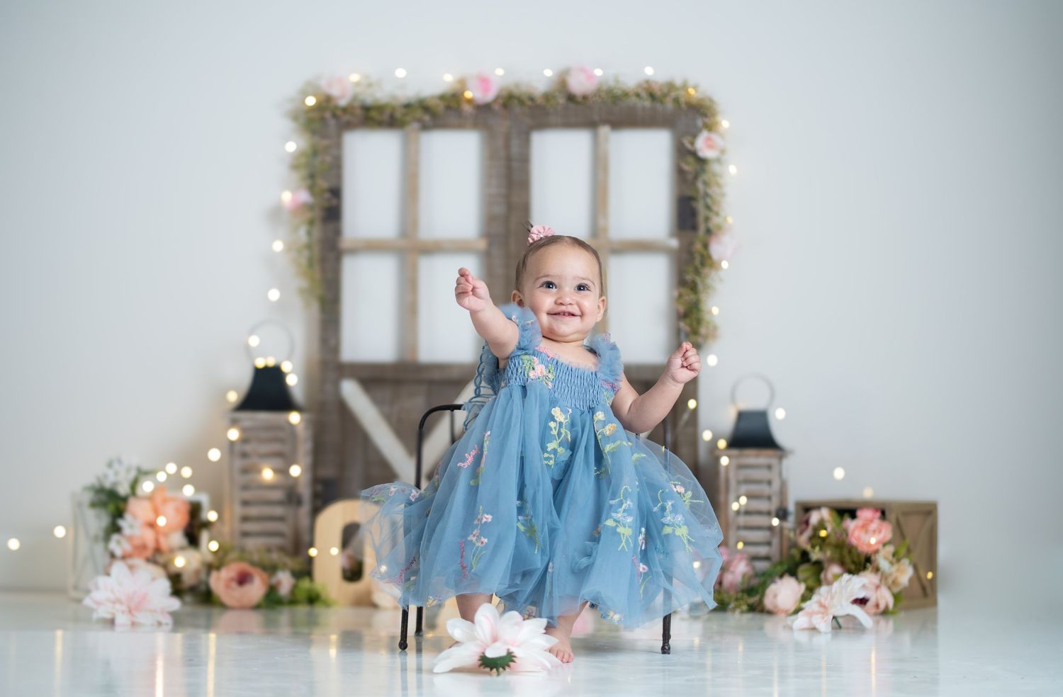 A smiling baby girl in a blue tulle dress sits among floral decorations and fairy lights in a professional photo shoot.