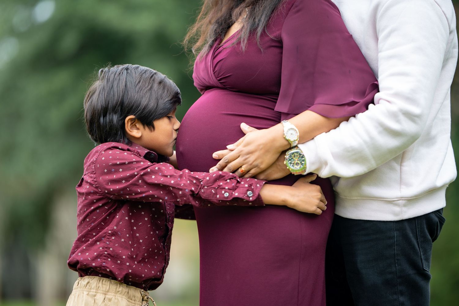A young child in a burgundy sweater touches the belly of a pregnant woman wearing a maroon dress.