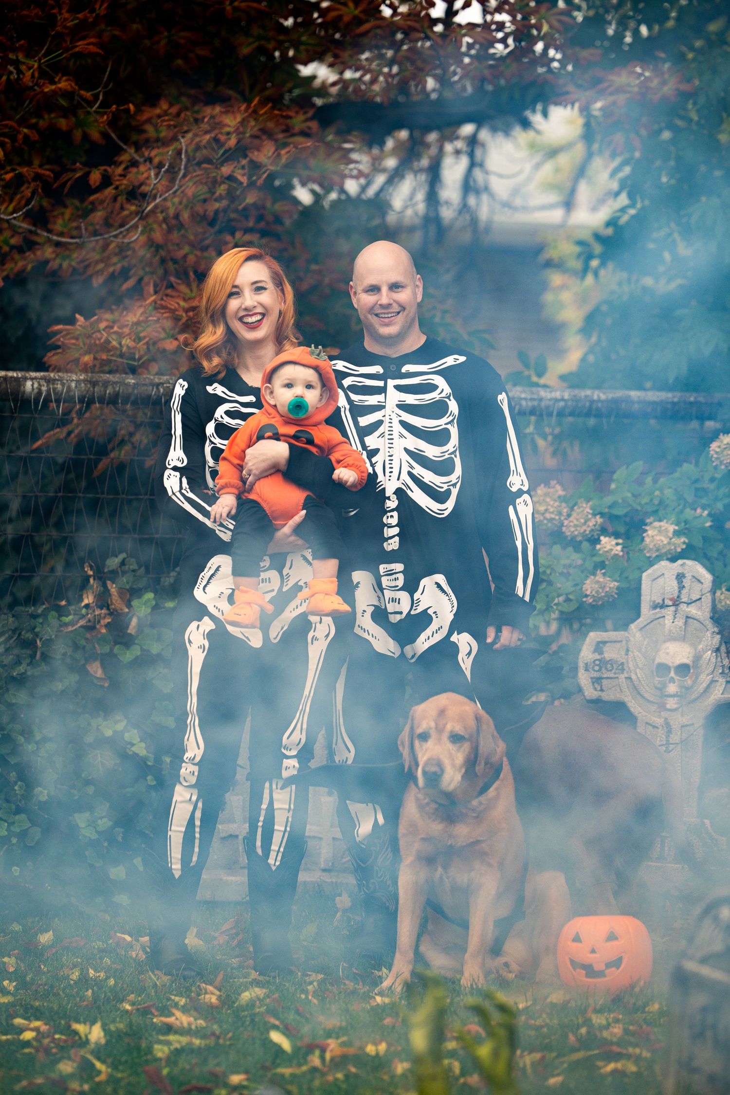 Family in skeleton costumes with dog and pumpkins in spooky Halloween scene.
