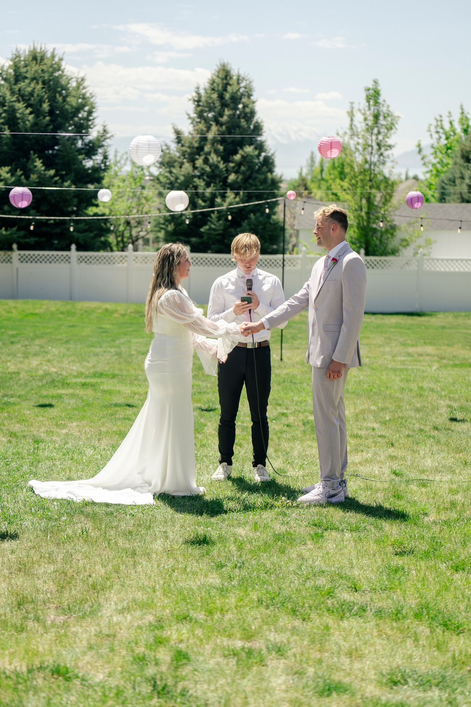 An outdoor wedding ceremony takes place on a sunny day with string lights overhead.