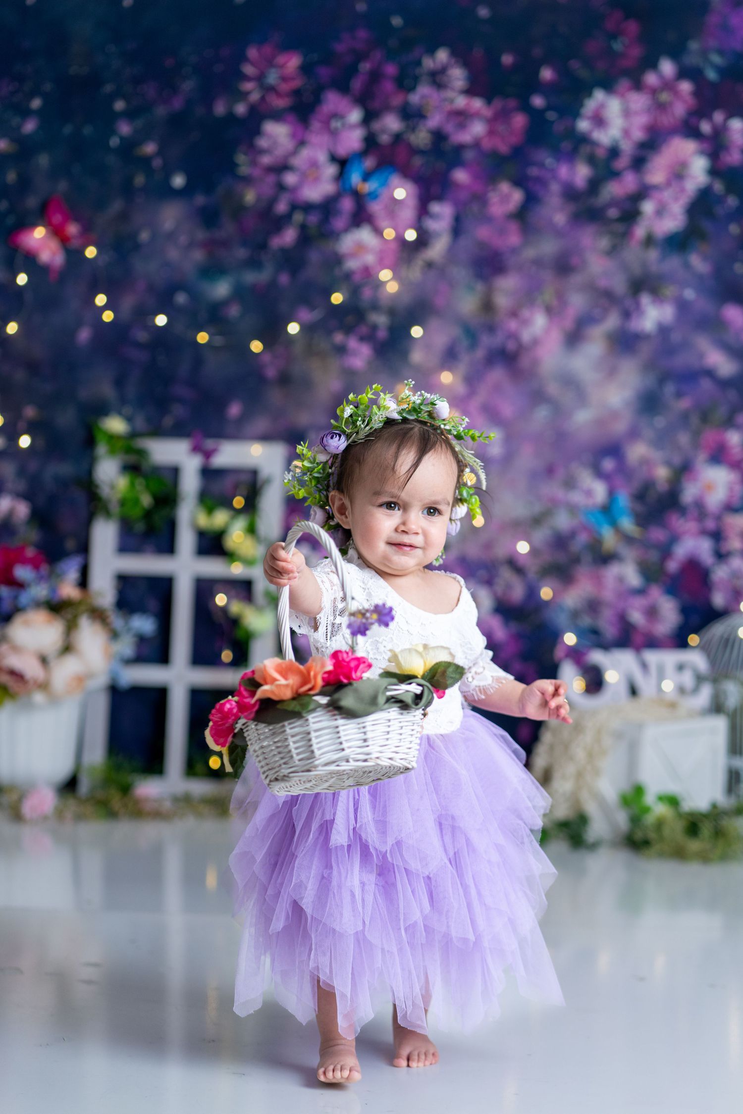 A child in a purple tulle dress and flower crown holds a white wicker basket against a floral backdrop.