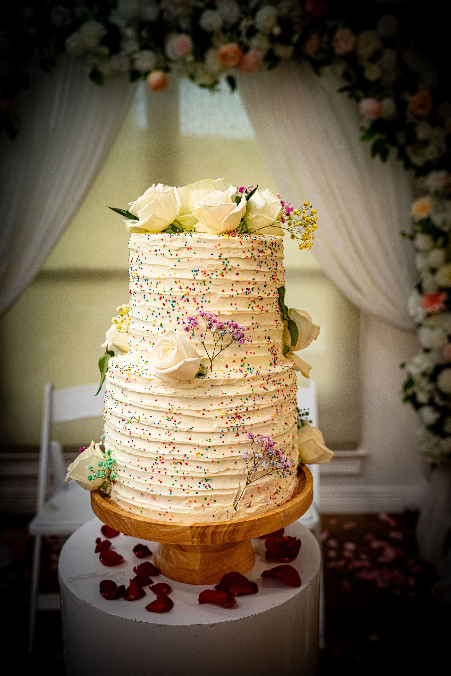 Rustic white wedding cake decorated with fresh flowers and textured frosting against draped backdrop.