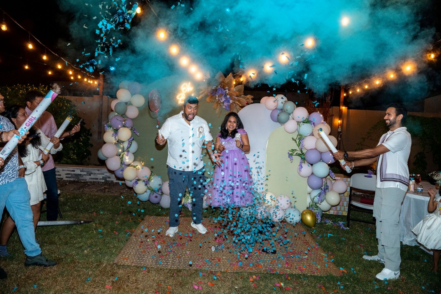 Outdoor celebration with balloon arch and blue smoke effects under string lights.